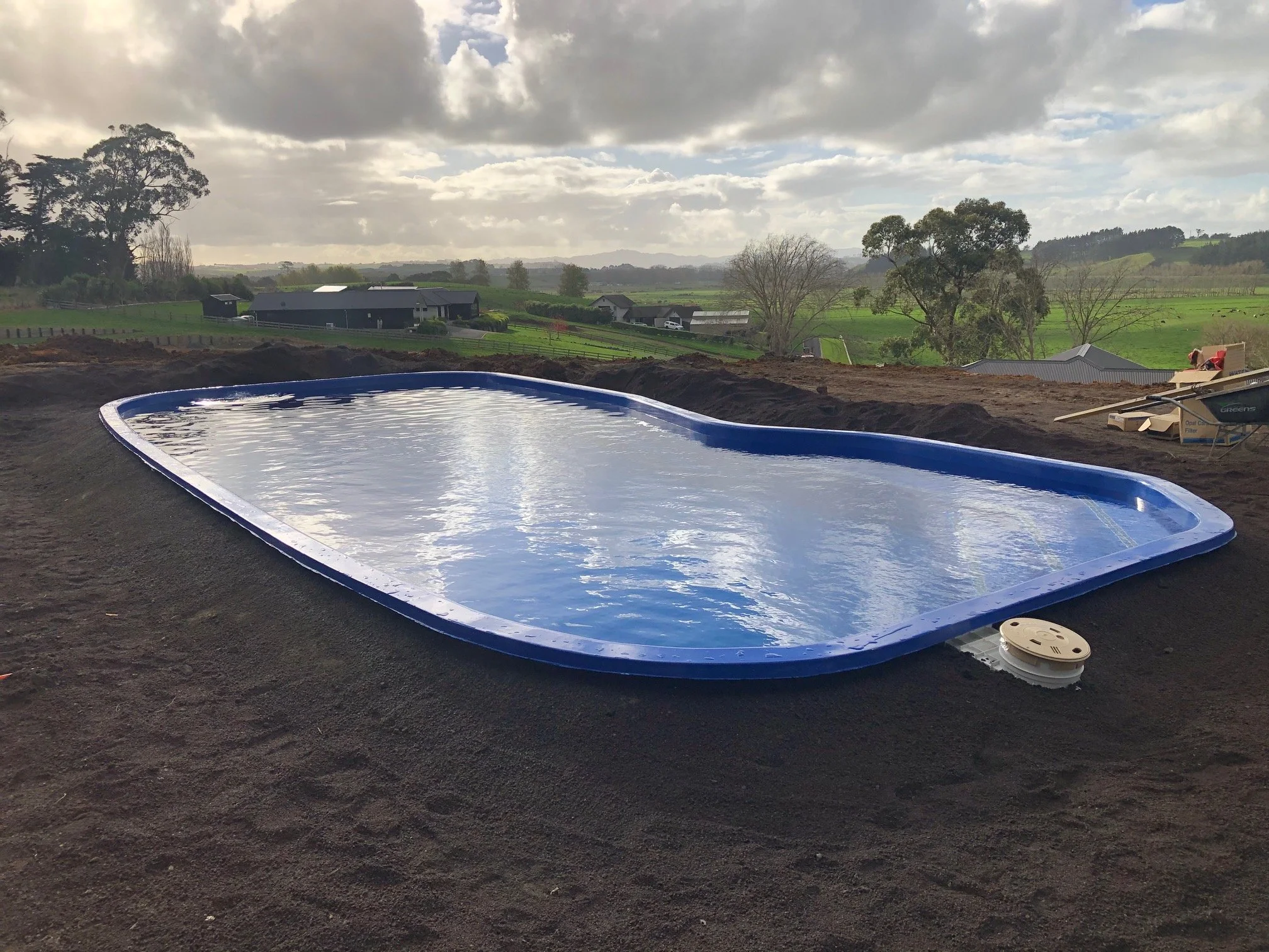 Inground swimming pool under construction in a rural area with green fields and trees in the background, and cloudy sky.