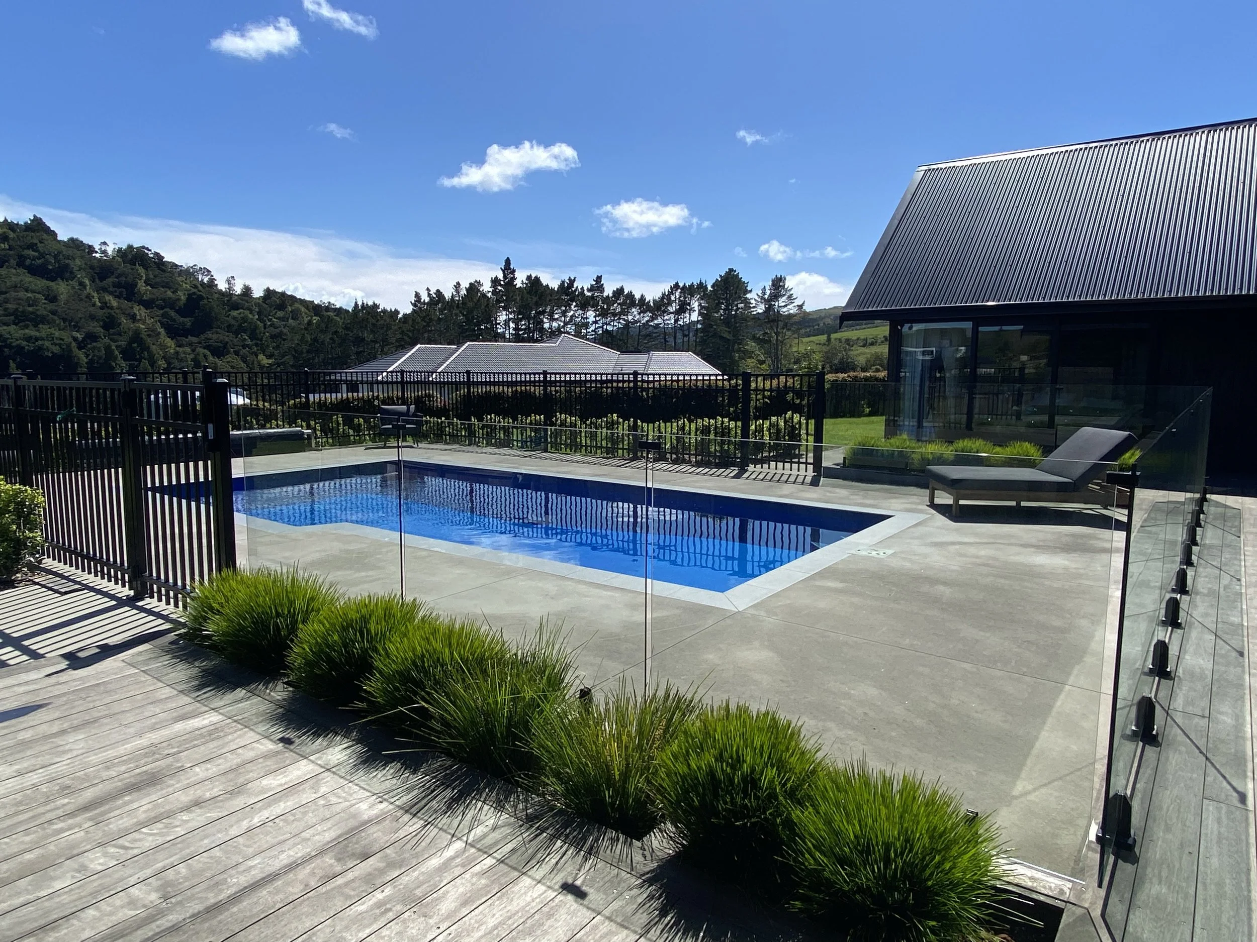A modern outdoor swimming pool surrounded by a glass fence, with a black lounge chair nearby, set in a landscaped backyard with green hills and trees in the background, under a blue sky with some clouds.