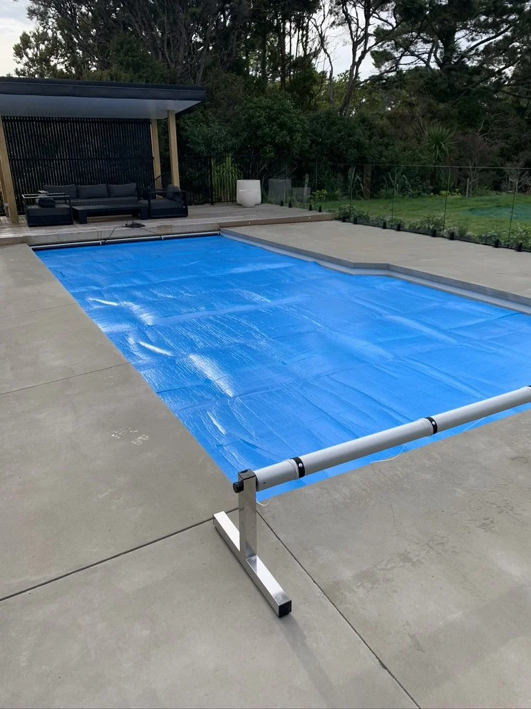 Empty backyard swimming pool covered with a blue pool cover, surrounded by concrete patio, with outdoor seating area in the background, greenery, and trees.
