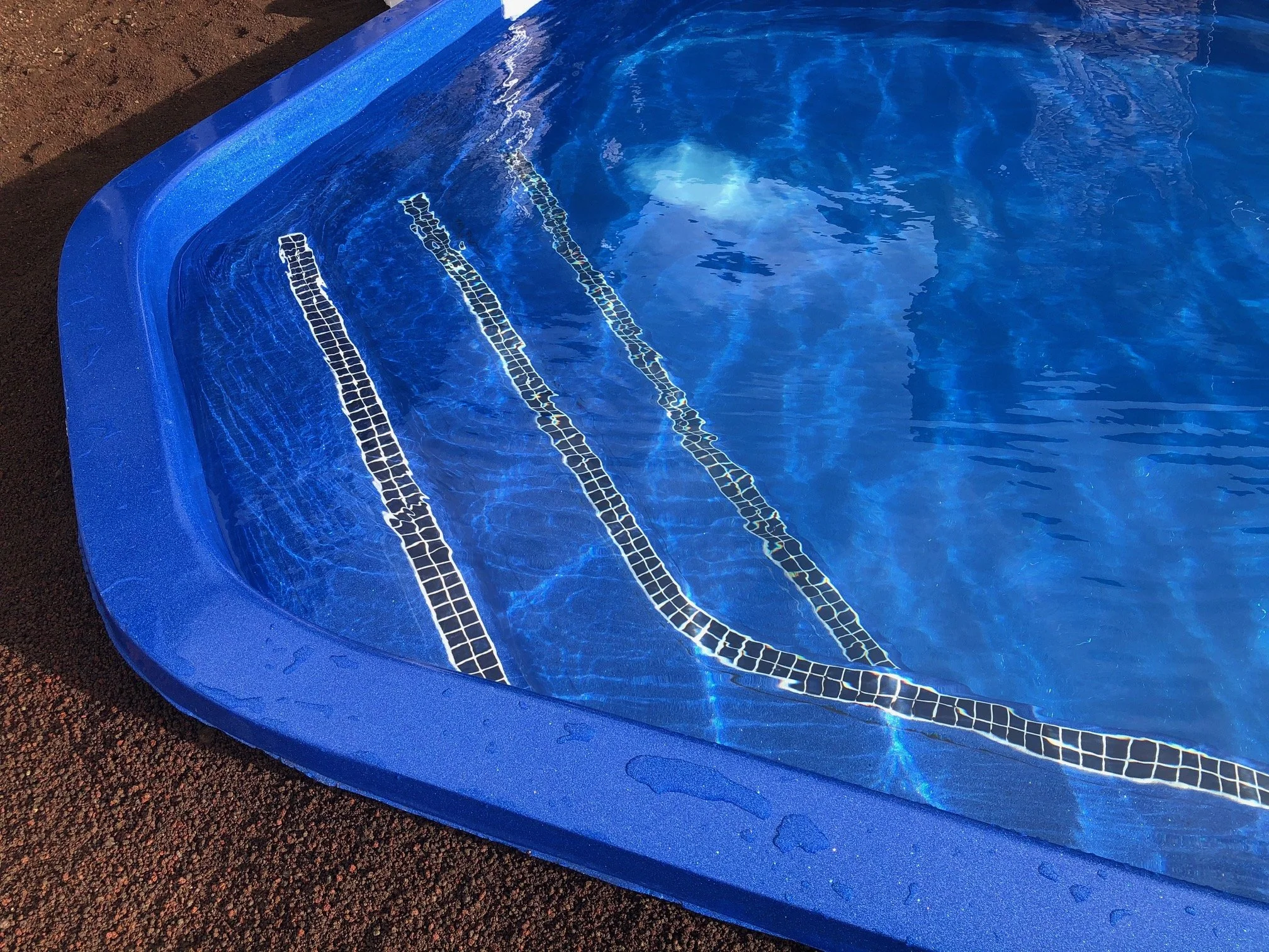 Close-up of a blue swimming pool with tiled stairs, water reflecting sunlight, and water droplets on the pool edge.