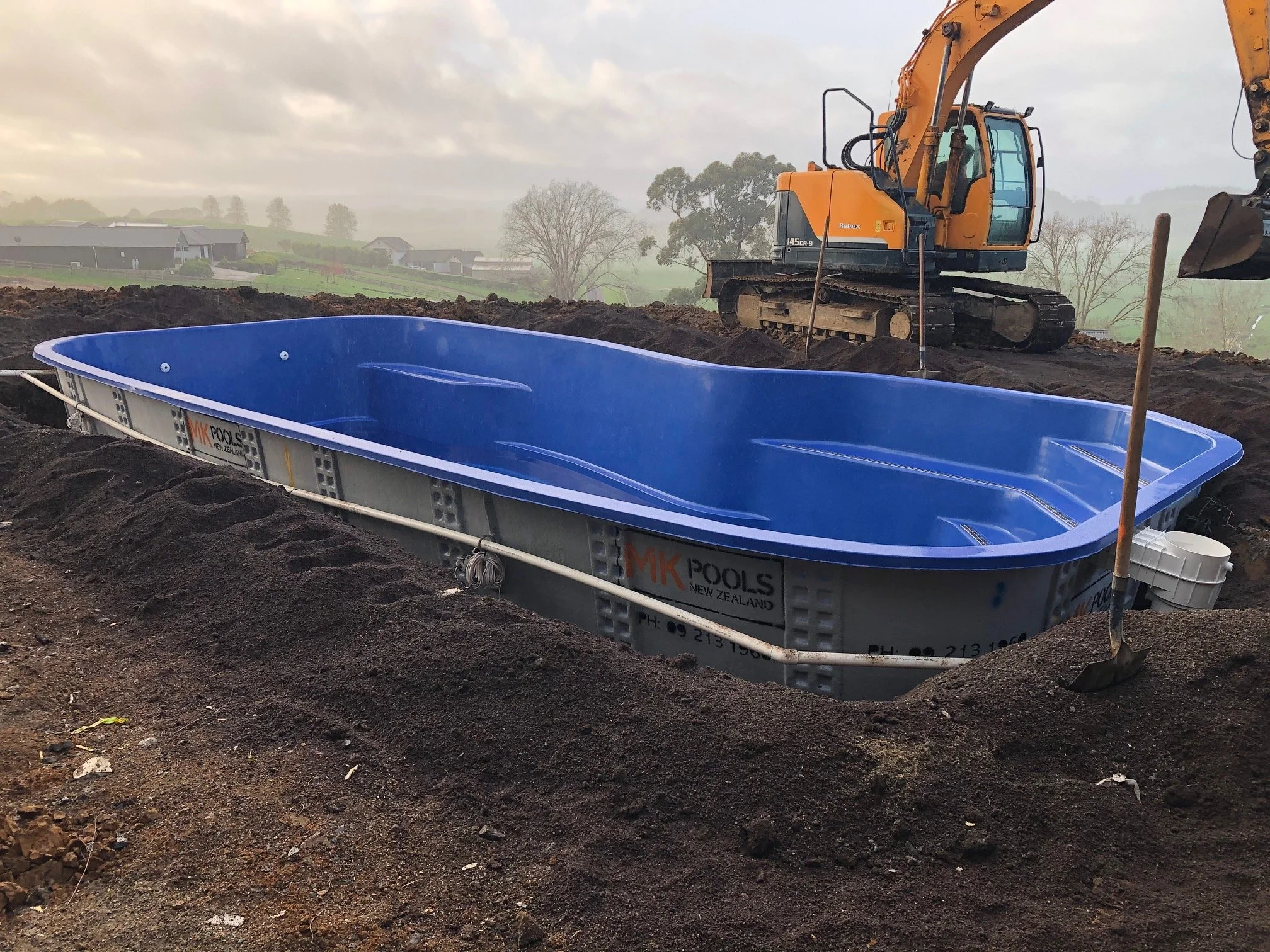 A blue swimming pool installed in a dug-out area with a construction excavator and shovels nearby, on a cloudy day in a rural area.