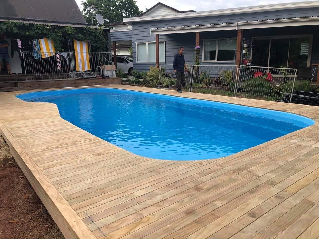 A backyard scene with a swimming pool surrounded by a wooden deck. There is a house with grey siding and a garden in the background. A man is standing near the fence that separates the pool area from the garden.