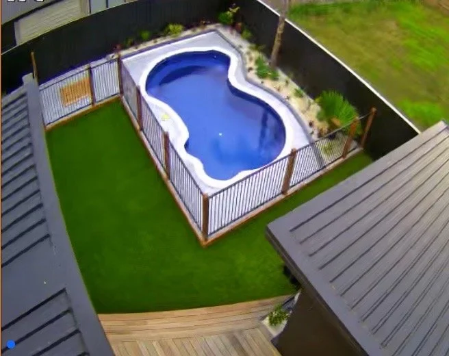 An above view of a backyard with a small kidney-shaped swimming pool surrounded by a wooden fence, grass lawn, and a deck with a gray roofed structure.