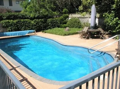 Swimming pool in a backyard with outdoor furniture, an umbrella, and lush greenery.