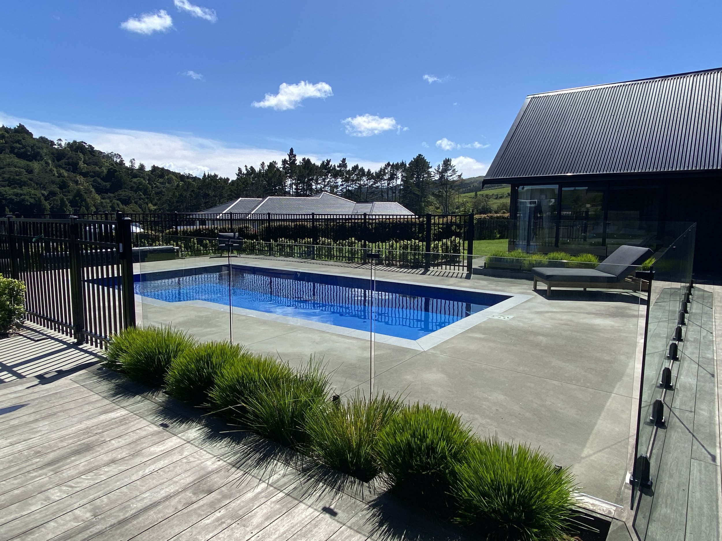 Modern backyard with a swimming pool, lounge chair, black metal fence, surrounding lush green landscape, blue sky with clouds, and adjacent black building with a metal roof.