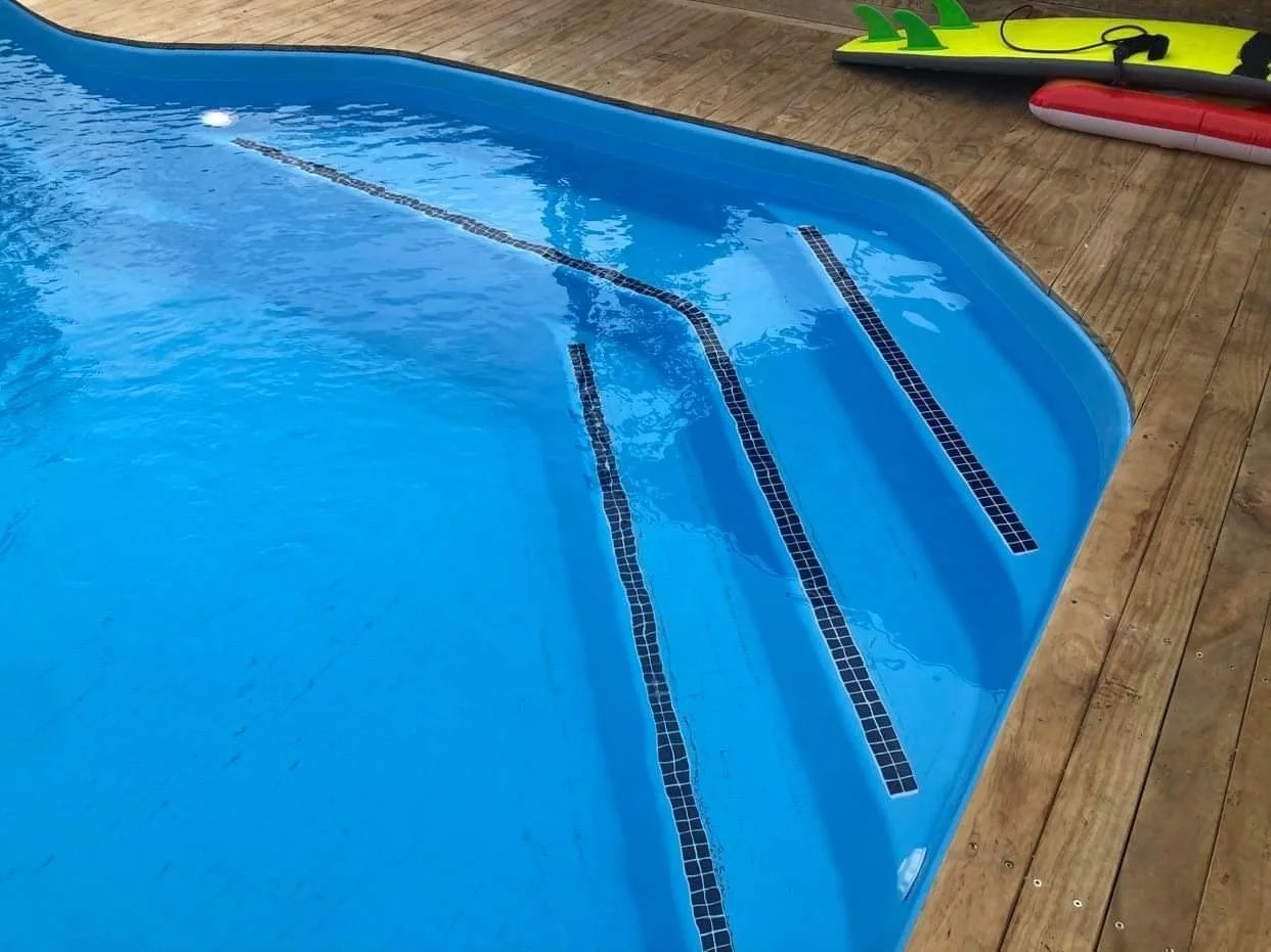 Close-up of a swimming pool with three blue tile lines on the bottom, next to a wooden deck. Surfboards and a paddleboard are lying on the deck.