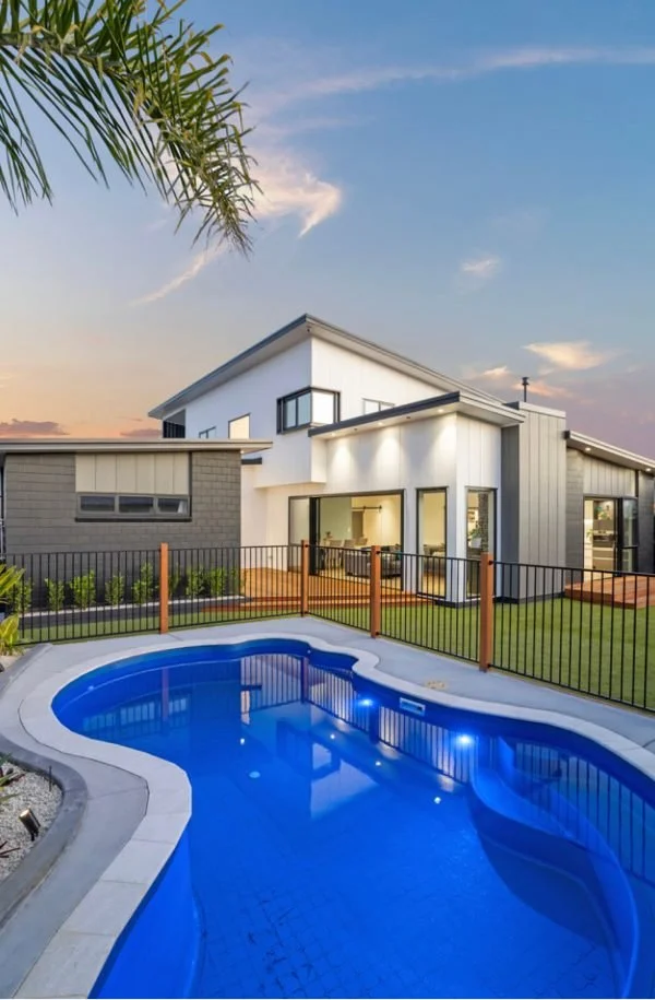 Modern house with large windows, a backyard with a pool, and a fenced yard during sunset.