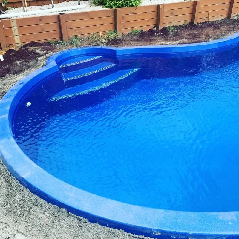 A blue above-ground swimming pool with built-in steps, located outdoors next to a wooden fence and green bushes.