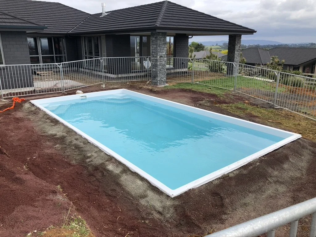 An in-ground rectangular swimming pool filled with clear blue water, surrounded by a patch of dirt, with a modern house in the background and a metal safety fence around the pool area.