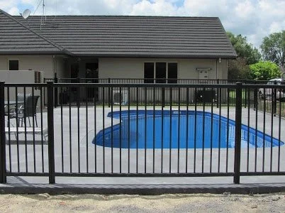 A backyard with a fenced-in in-ground swimming pool, patio chairs, and a house in the background.