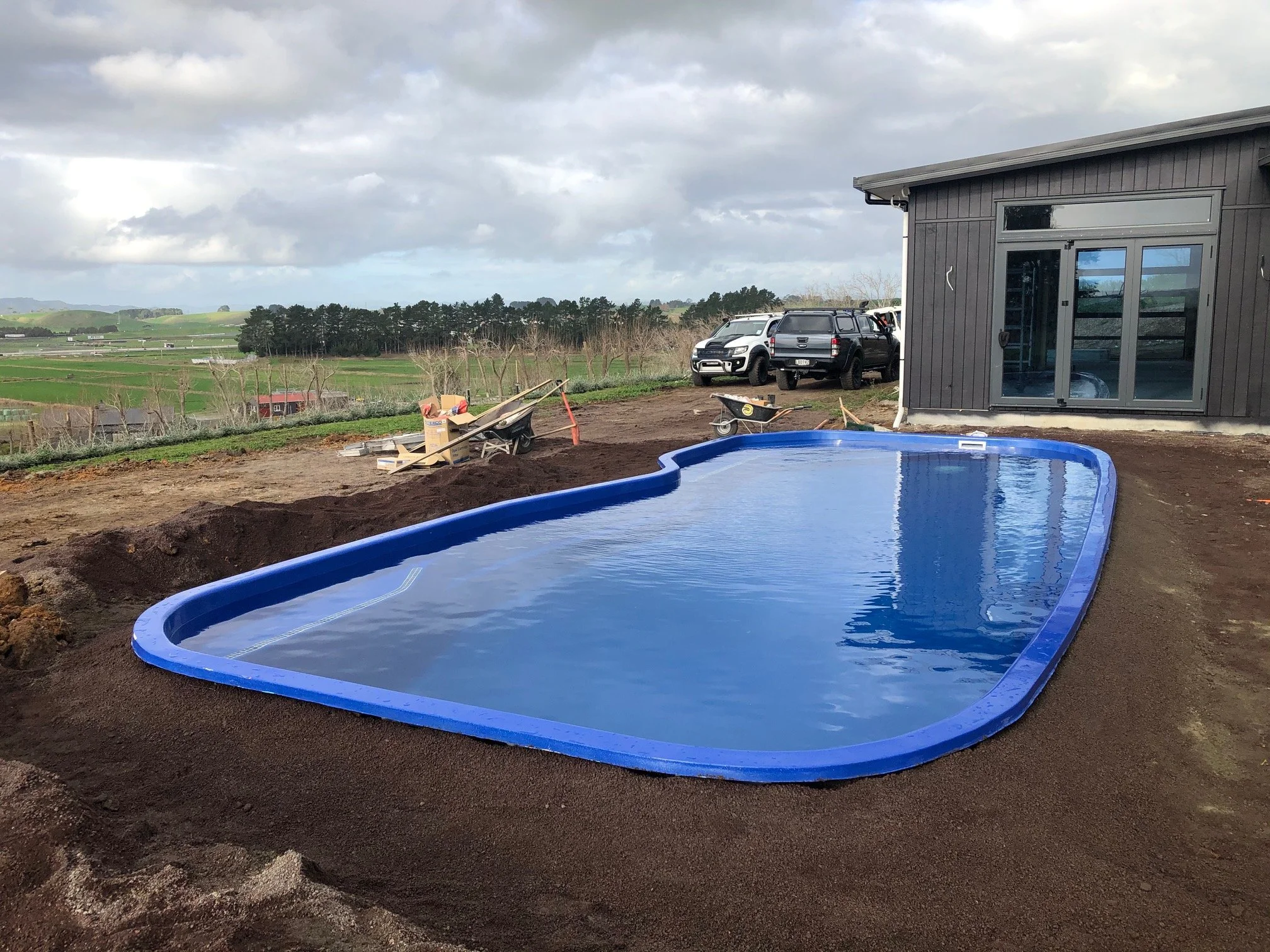 Empty in-ground swimming pool with a blue lining partially filled with water, next to a black house with large sliding glass doors, on a dirt yard. Vehicles parked nearby and construction tools around.