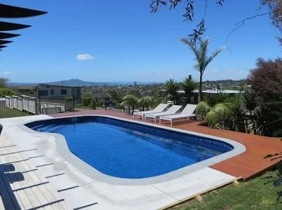 Swimming pool in a backyard with lounge chairs, trees, and a city view in the distance.