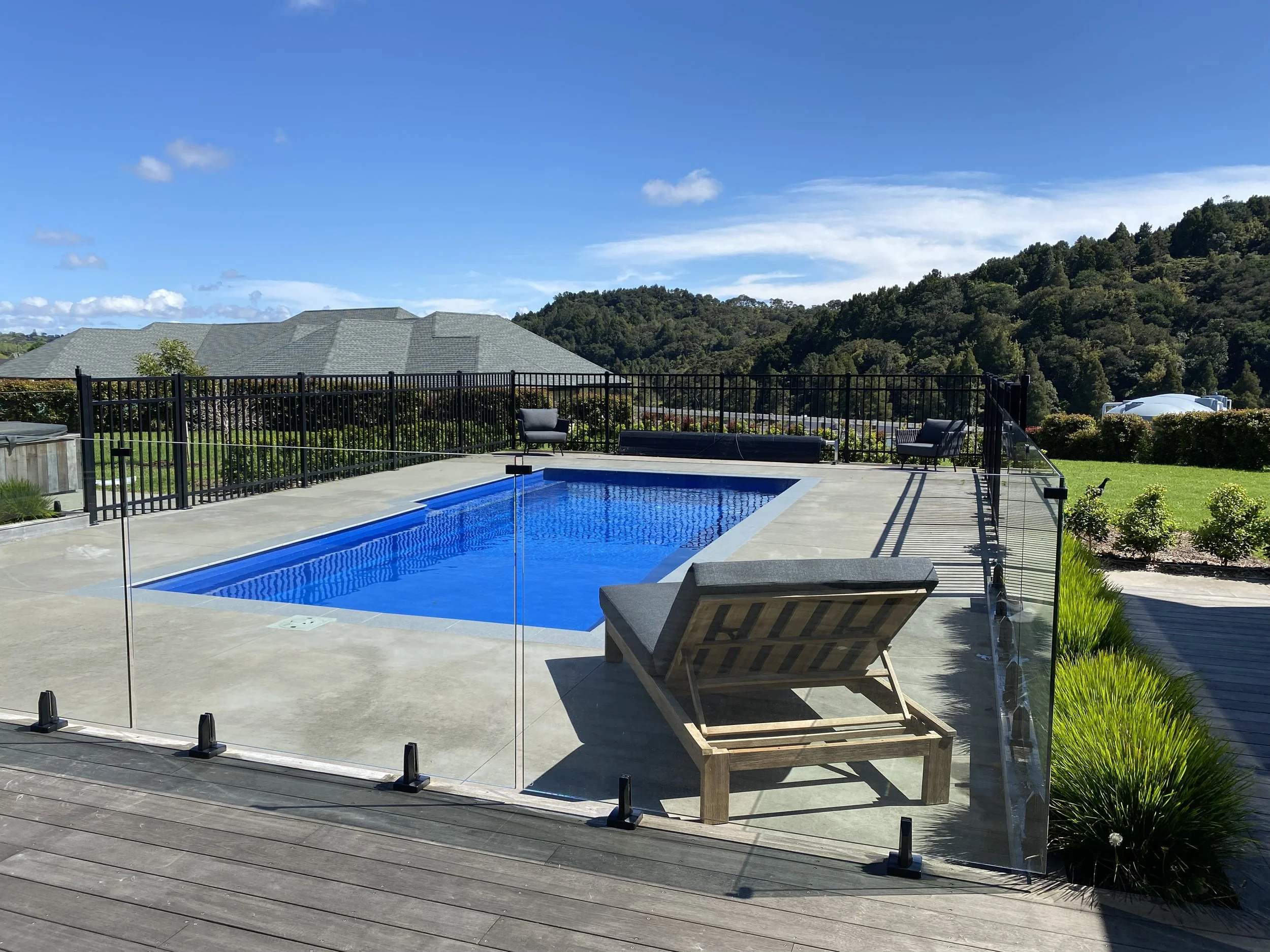 A backyard swimming pool with a clear glass fence, surrounded by lounge chairs, greenery, and hilly landscape in the background under a blue sky with some clouds.