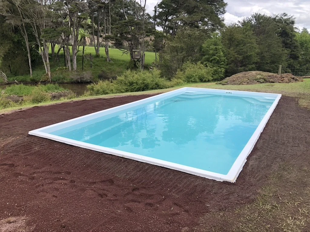 An empty rectangular pool filled with blue water, surrounded by dirt and green grass with trees and bushes in the background.
