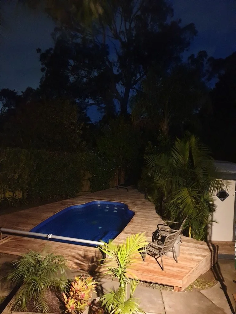 Nighttime backyard scene with a small swimming pool on a wooden deck, surrounded by lush greenery and tall trees.