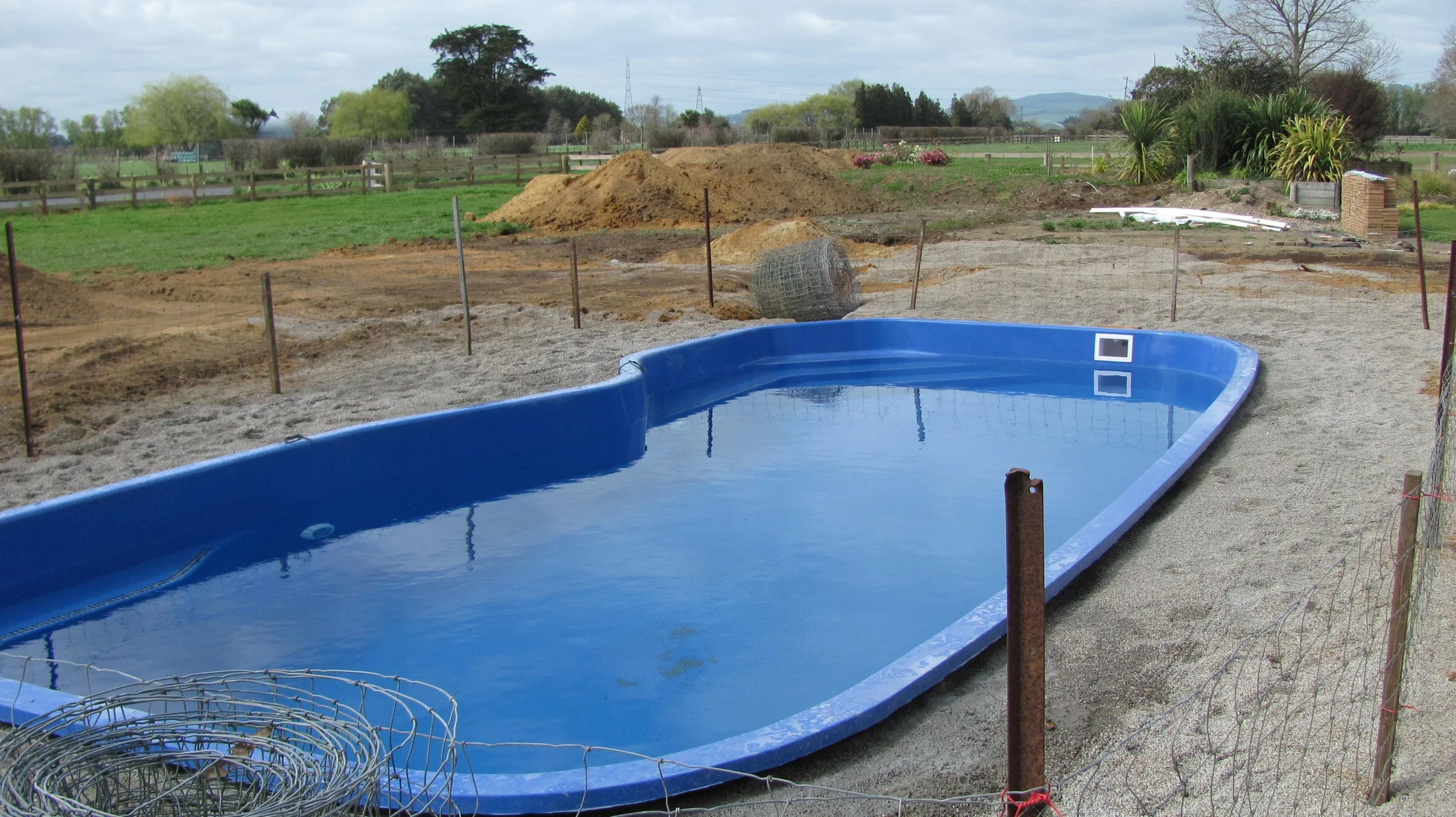 An outdoor construction site with a blue swimming pool filled with water, surrounded by sandy and dirt areas, with piles of dirt and construction materials in the background, and a rural landscape with trees and hills in the distance.