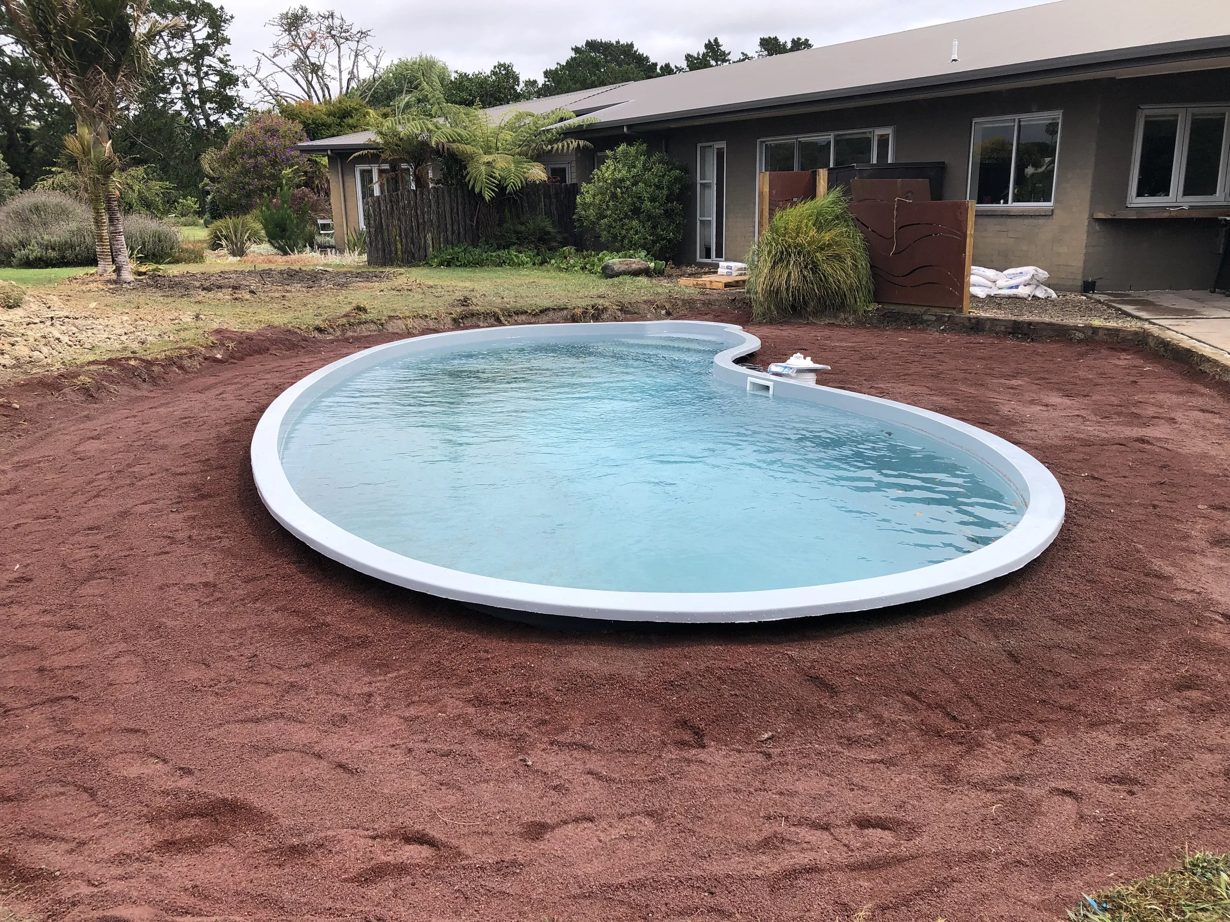 In-ground kidney-shaped swimming pool in the backyard with red soil surrounding it, next to a house with a deck and a variety of plants and trees.