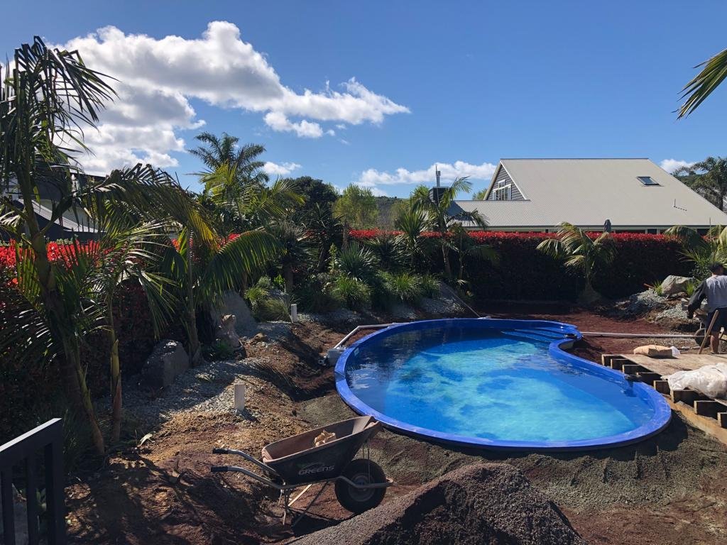 A backyard with a small kidney-shaped swimming pool under construction, surrounded by tropical plants, with a house in the background and a person working on the construction.
