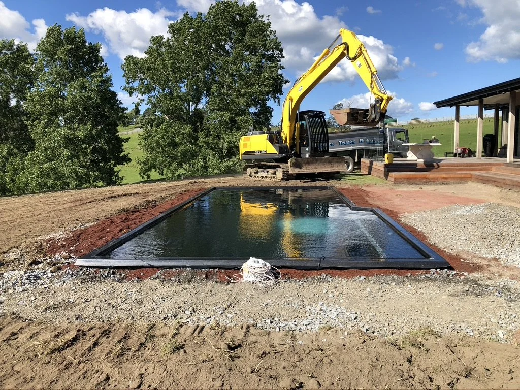A construction site showing a small excavator with a yellow arm, filling a black frame pool with water. The site is outdoors on a sunny day with green trees and a blue sky with clouds in the background.