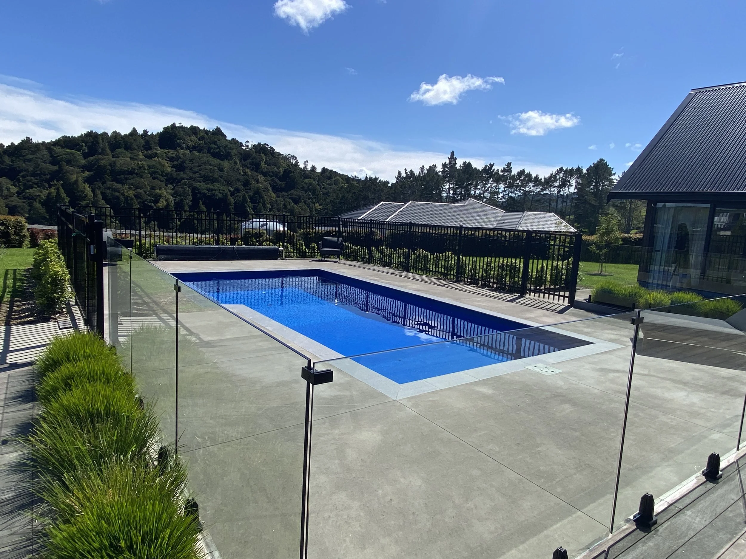 A modern backyard with a rectangular swimming pool surrounded by a glass fence, with a hillside and trees in the background under a blue sky with clouds.