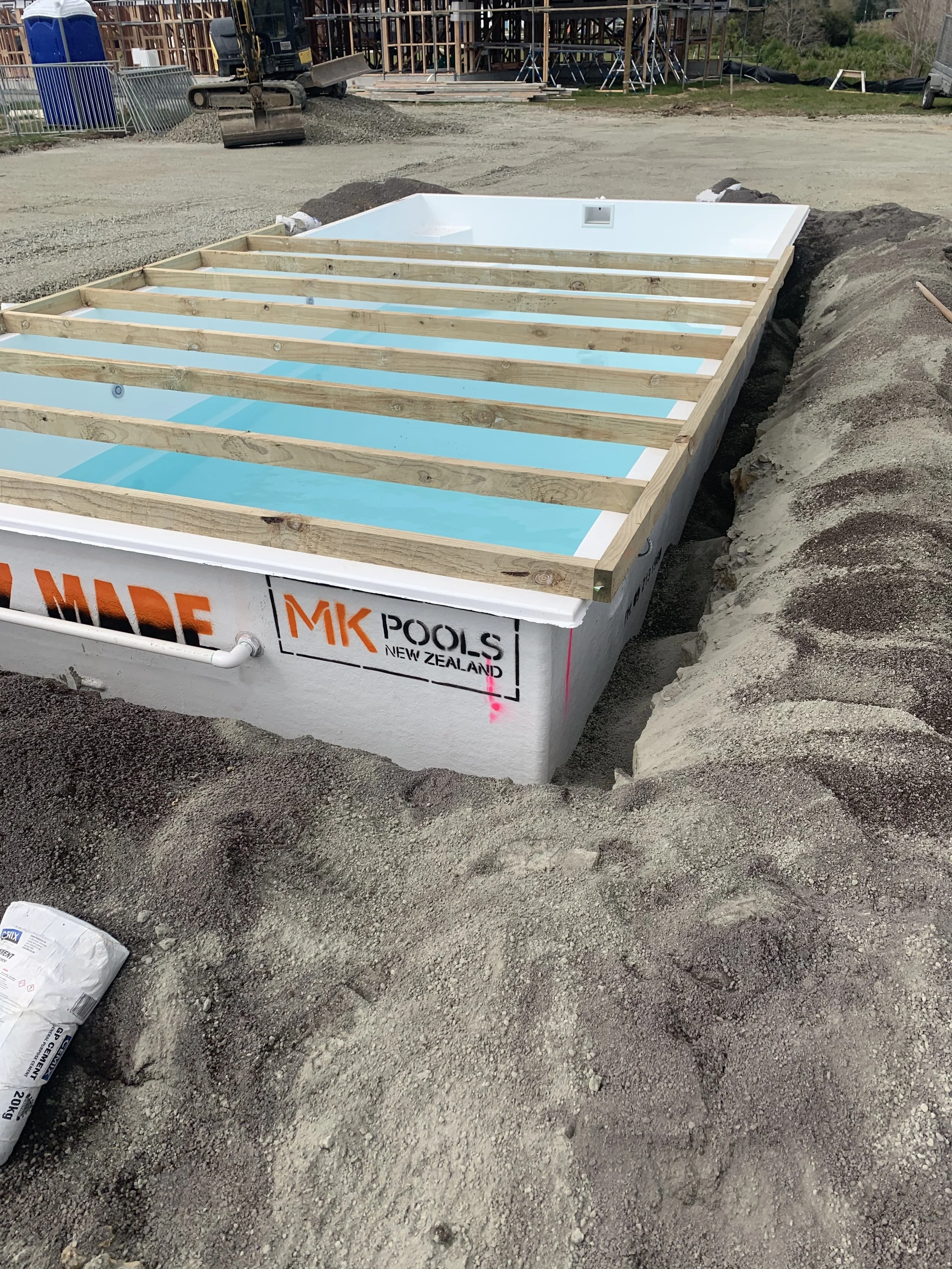 Swimming pool under construction with wooden beams, surrounded by dirt and construction materials at a construction site.