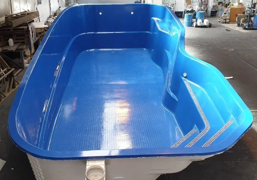 A large, blue plastic swimming pool with a textured interior, placed in an industrial warehouse with other construction and manufacturing materials in the background.