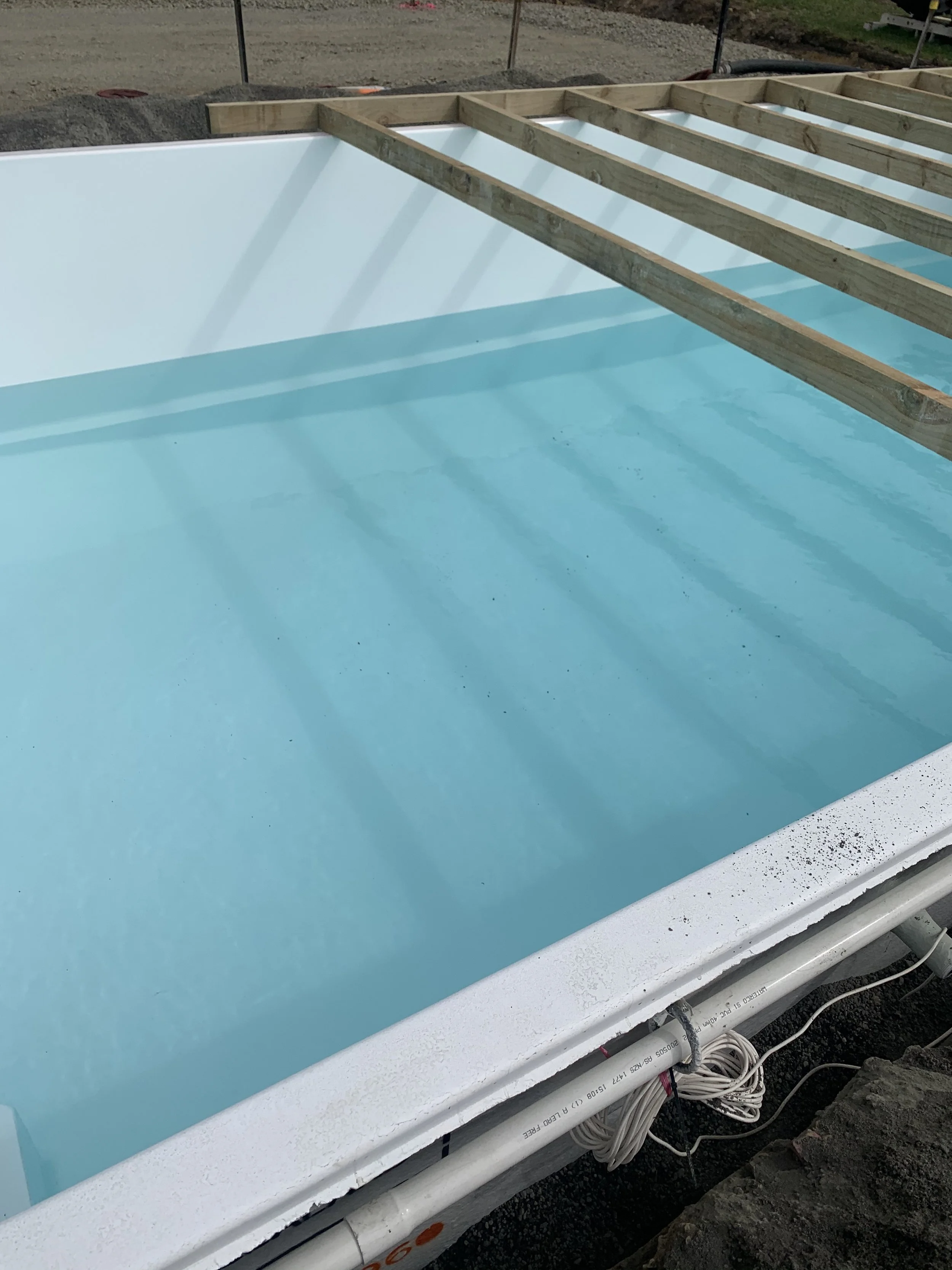 A partially constructed swimming pool with a white interior and light blue water, with wooden framing around it and visible pipes underneath.