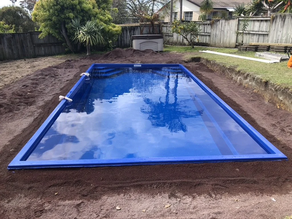 A rectangular, above-ground swimming pool with a blue frame filled with water, situated on a dirt surface in a backyard with trees, grass, and wooden fences.