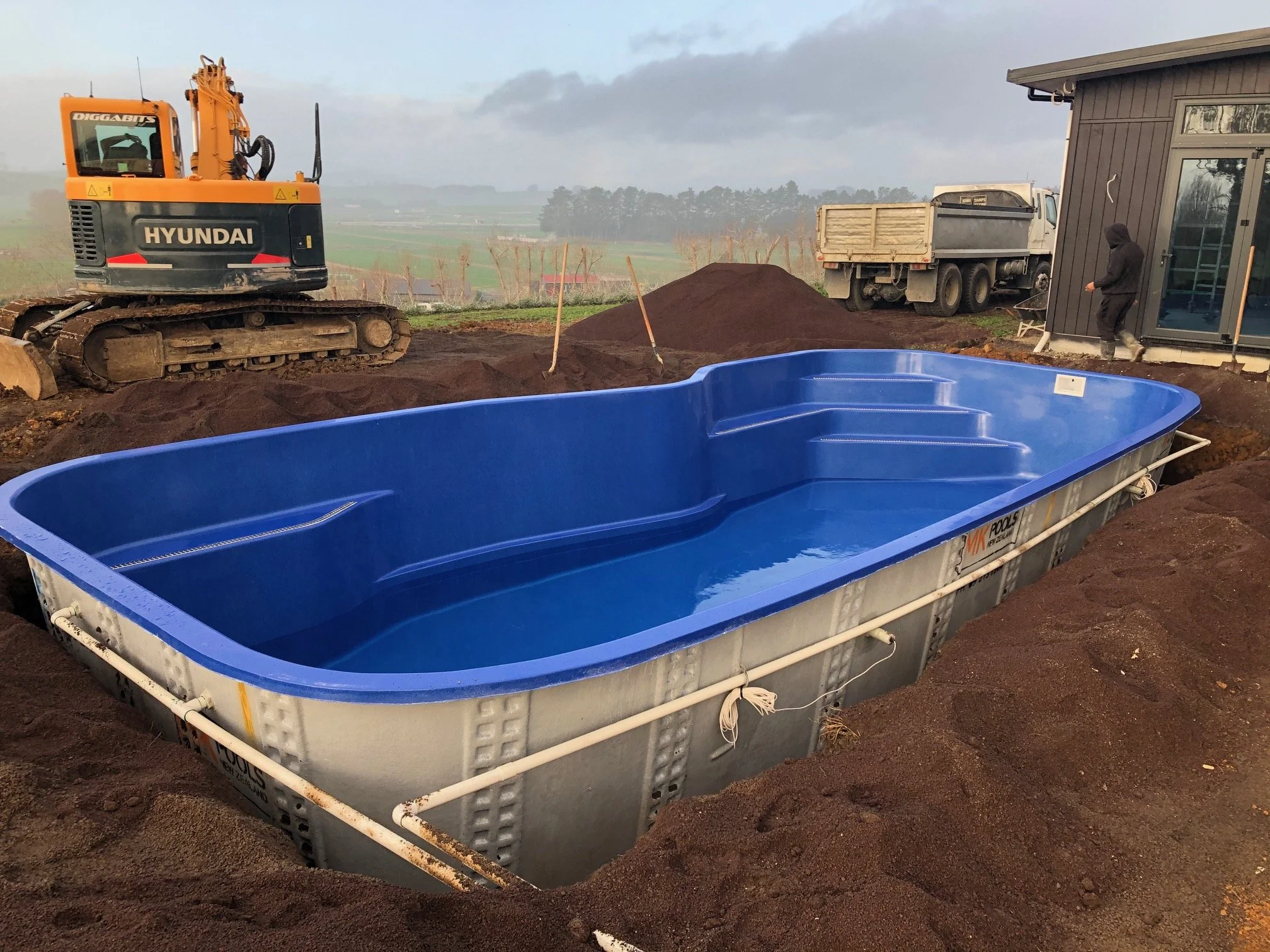 A large, empty, blue swimming pool installed in a dirt excavation site with a construction excavator, trucks, and a person nearby, on a cloudy day.