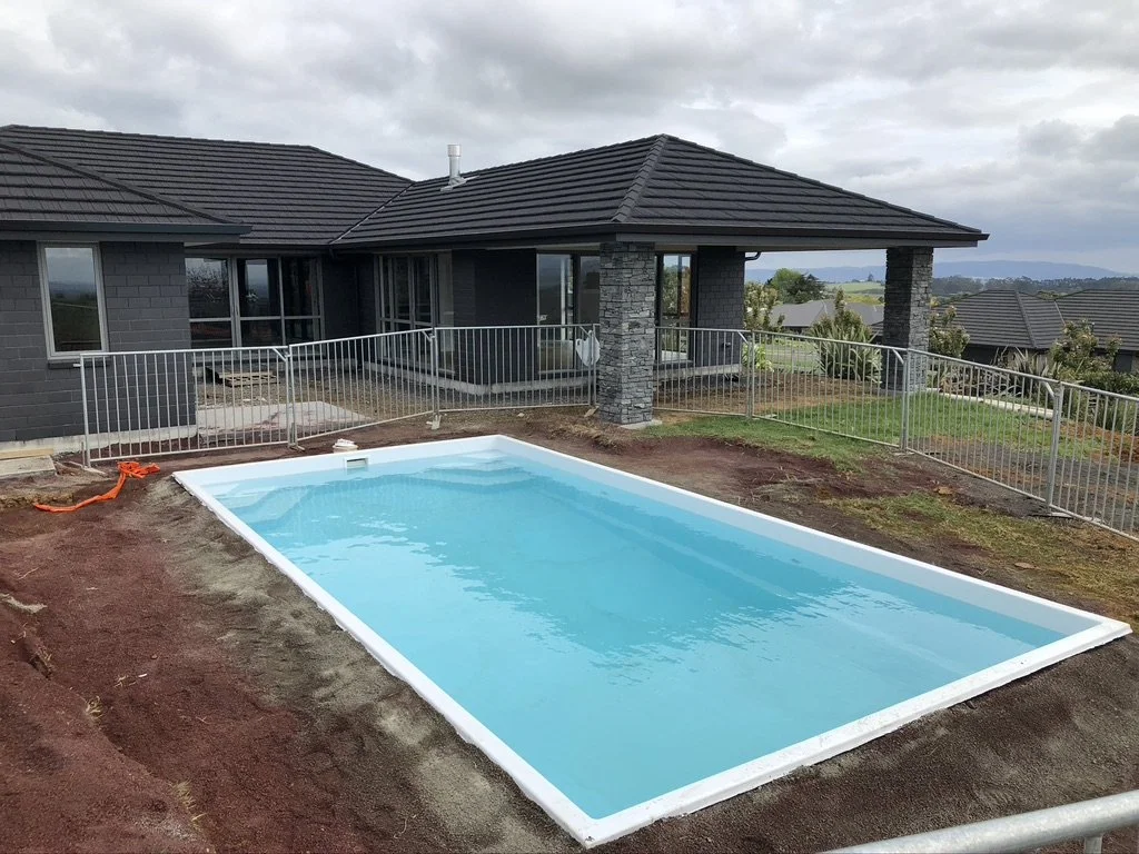 A backyard with a small in-ground swimming pool surrounded by a metal fence, a covered porch, and a gray house with a dark roof under a cloudy sky.