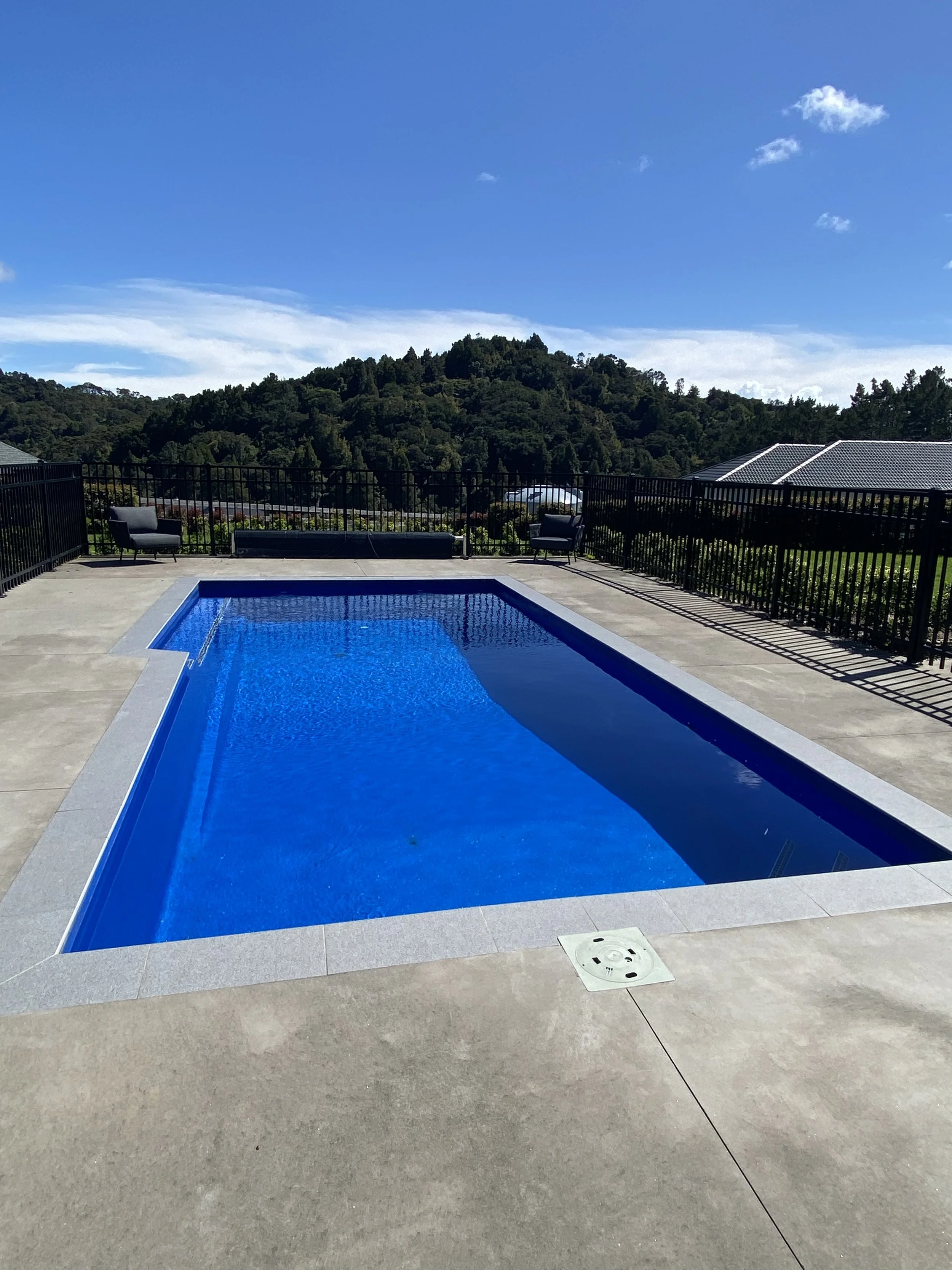 An outdoor swimming pool with a blue interior, surrounded by a concrete patio, with a mountainous landscape and blue sky in the background.