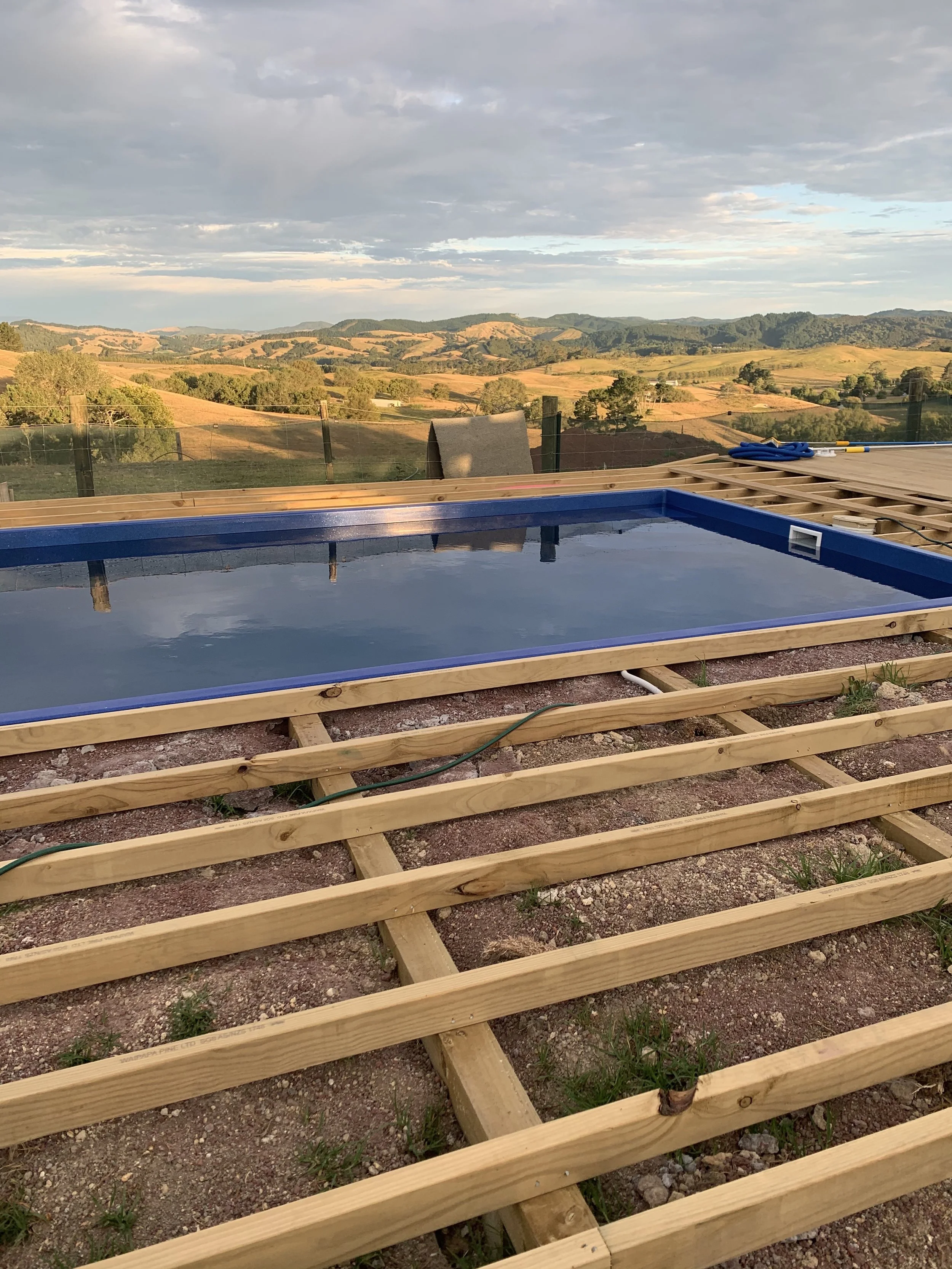 Construction site with a swimming pool in progress, wooden framework and a scenic landscape of rolling hills and a cloudy sky in the background.