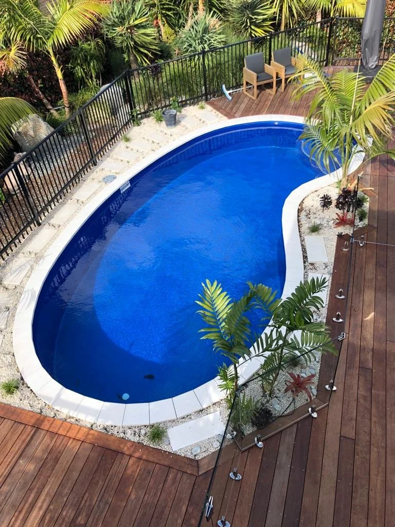 A backyard swimming pool with a curved shape, surrounded by a wooden deck and tropical plants. There are two chairs and a small table on a nearby patio area.