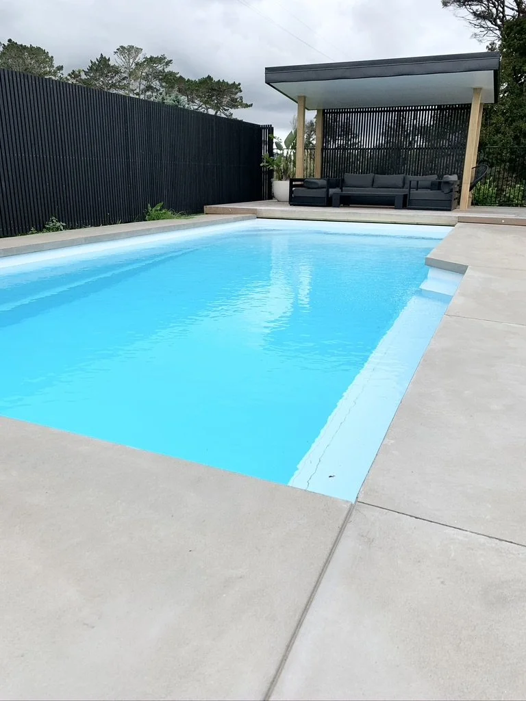 A modern backyard pool with light blue water, concrete decking, and a covered patio area with black outdoor furniture and tall potted plants.