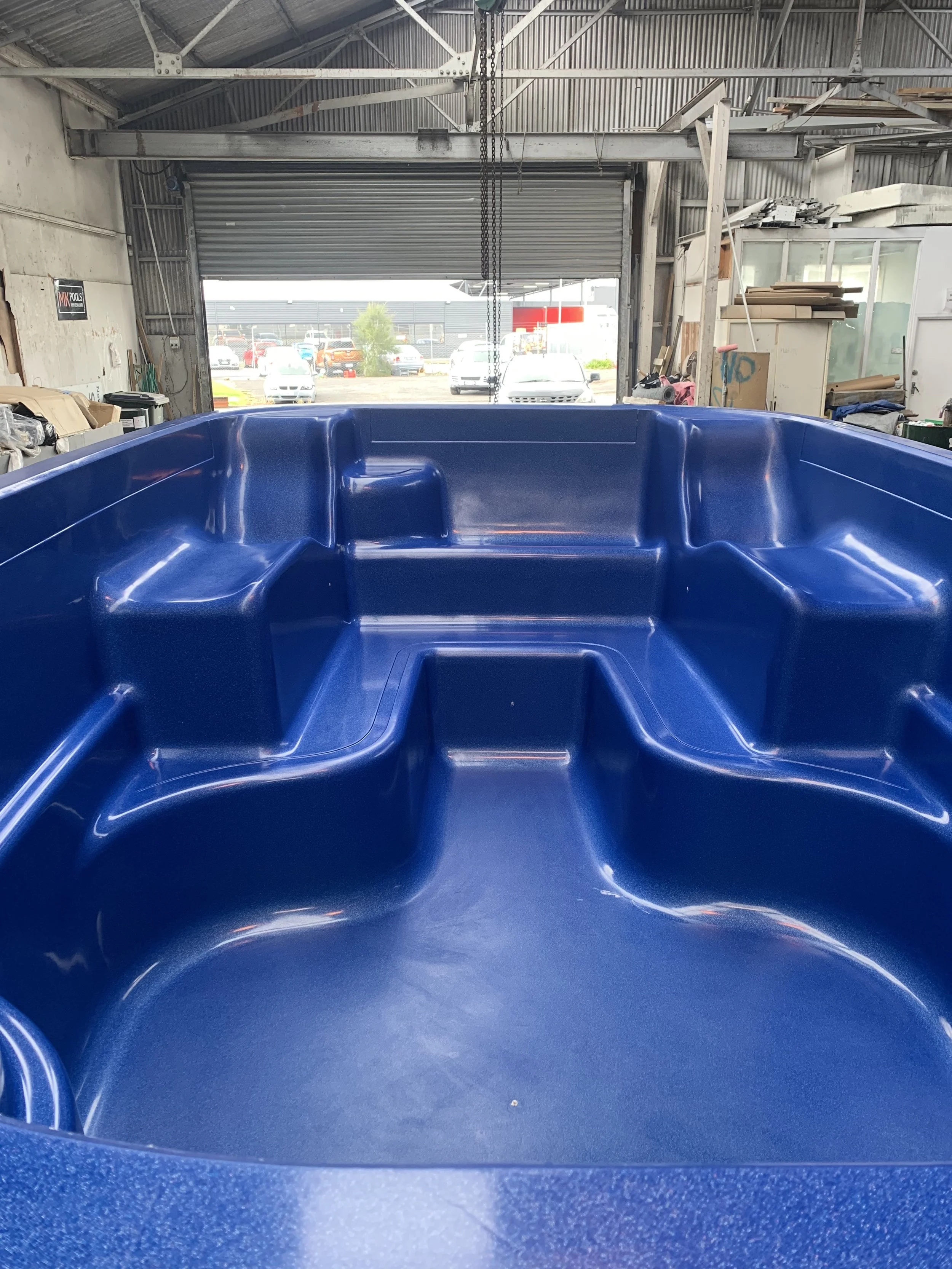 The image shows the interior of a large blue hot tub or spa with built-in seating and a textured surface, inside a garage or warehouse with an open garage door.
