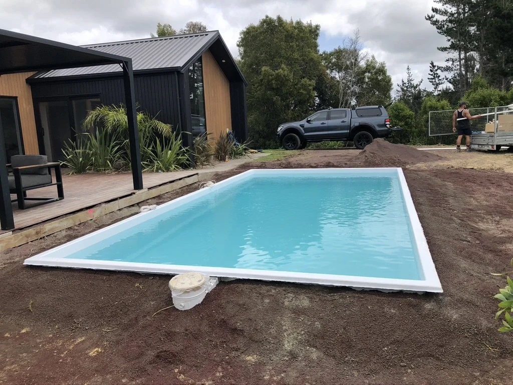 A newly installed above-ground swimming pool filled with water is situated on bare soil in a backyard, near a modern house with a wooden and black exterior, and some surrounding trees. A person is seen near a trailer with boxes, and a pickup truck is