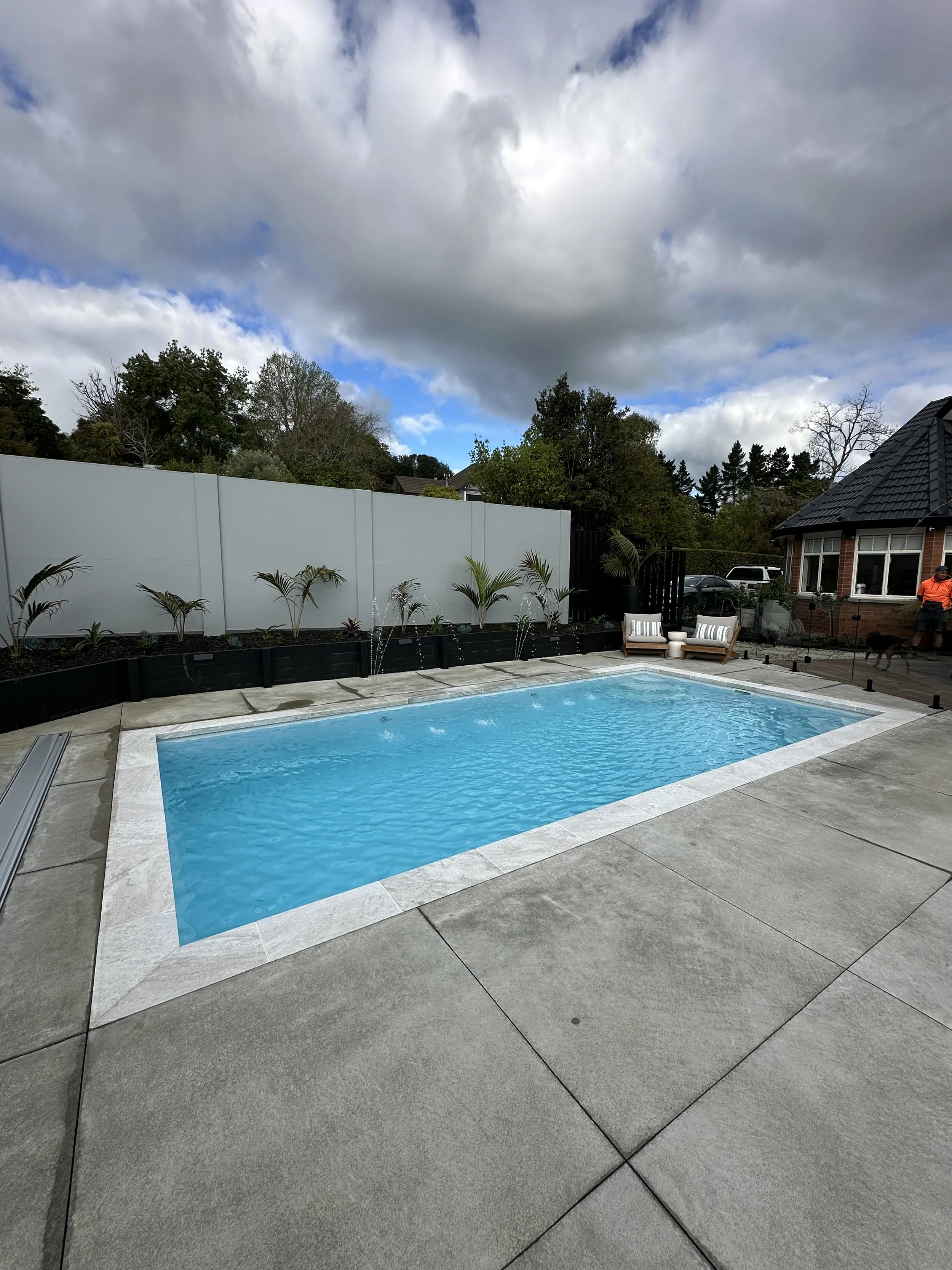 Residential backyard with a small swimming pool, decorative plants in black planters, and outdoor seating, under a cloudy sky.