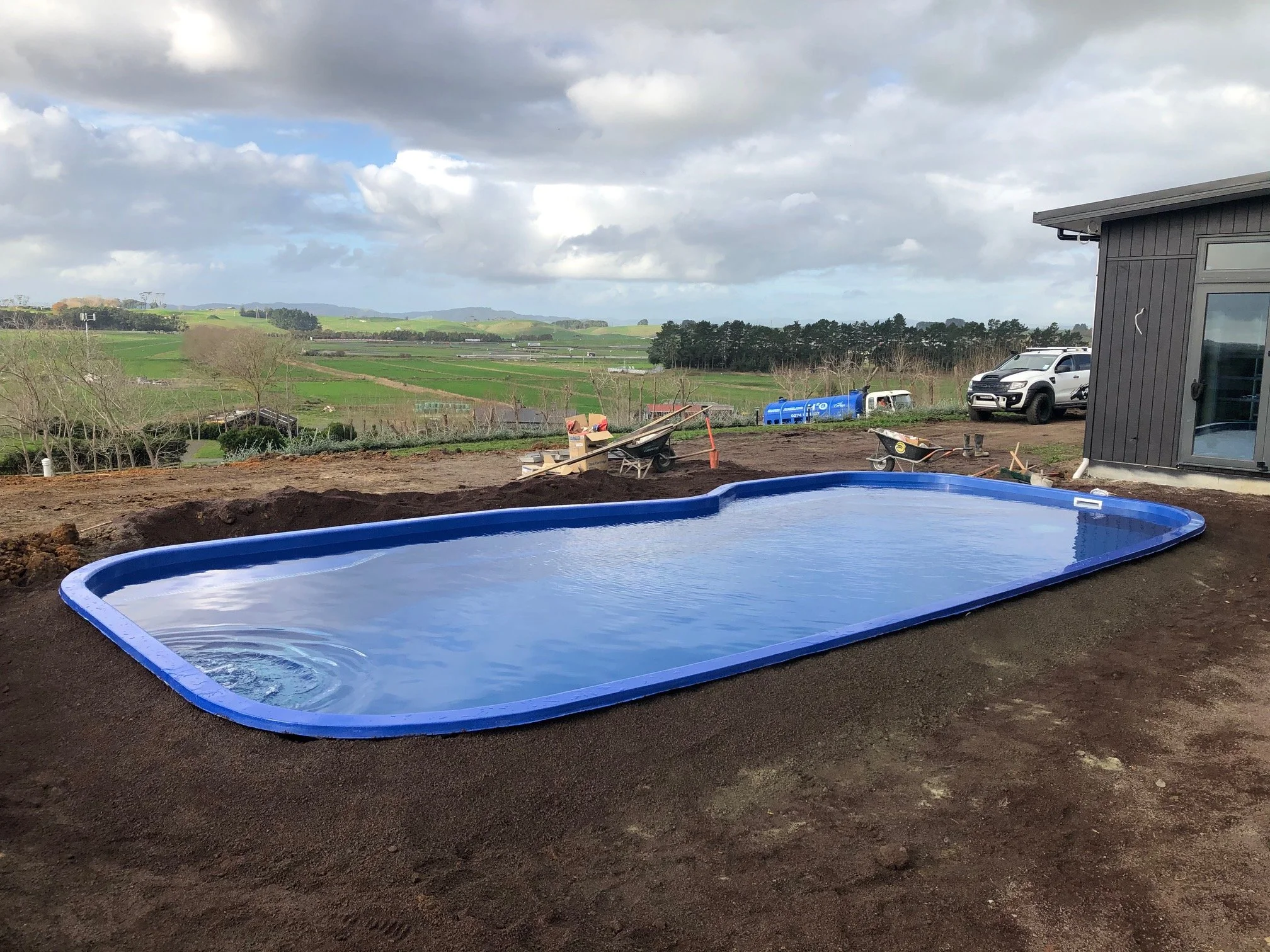 Uninstalled blue swimming pool in a backyard with a view of fields and hills, next to a gray house with large windows, construction tools and vehicles nearby.