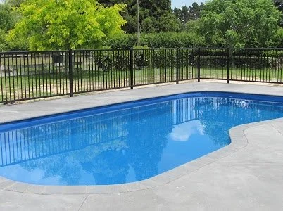 Empty swimming pool with a black metal fence surrounding it, green trees in the background, and a cloudy reflection on the water.