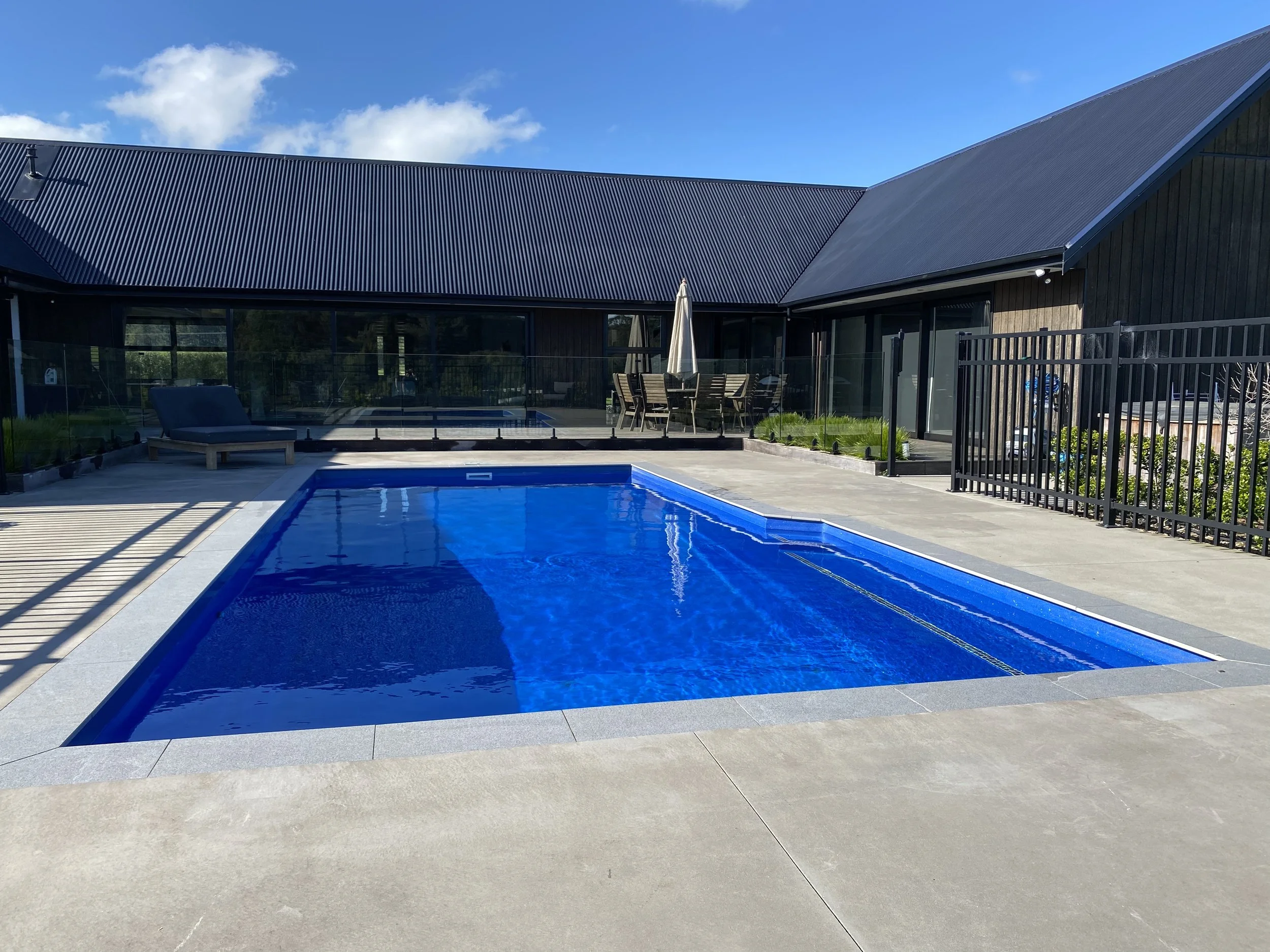 A modern backyard with a blue swimming pool, a lounge chair, a patio table with an umbrella, and a house with dark siding and a metal roof under a partly cloudy sky.