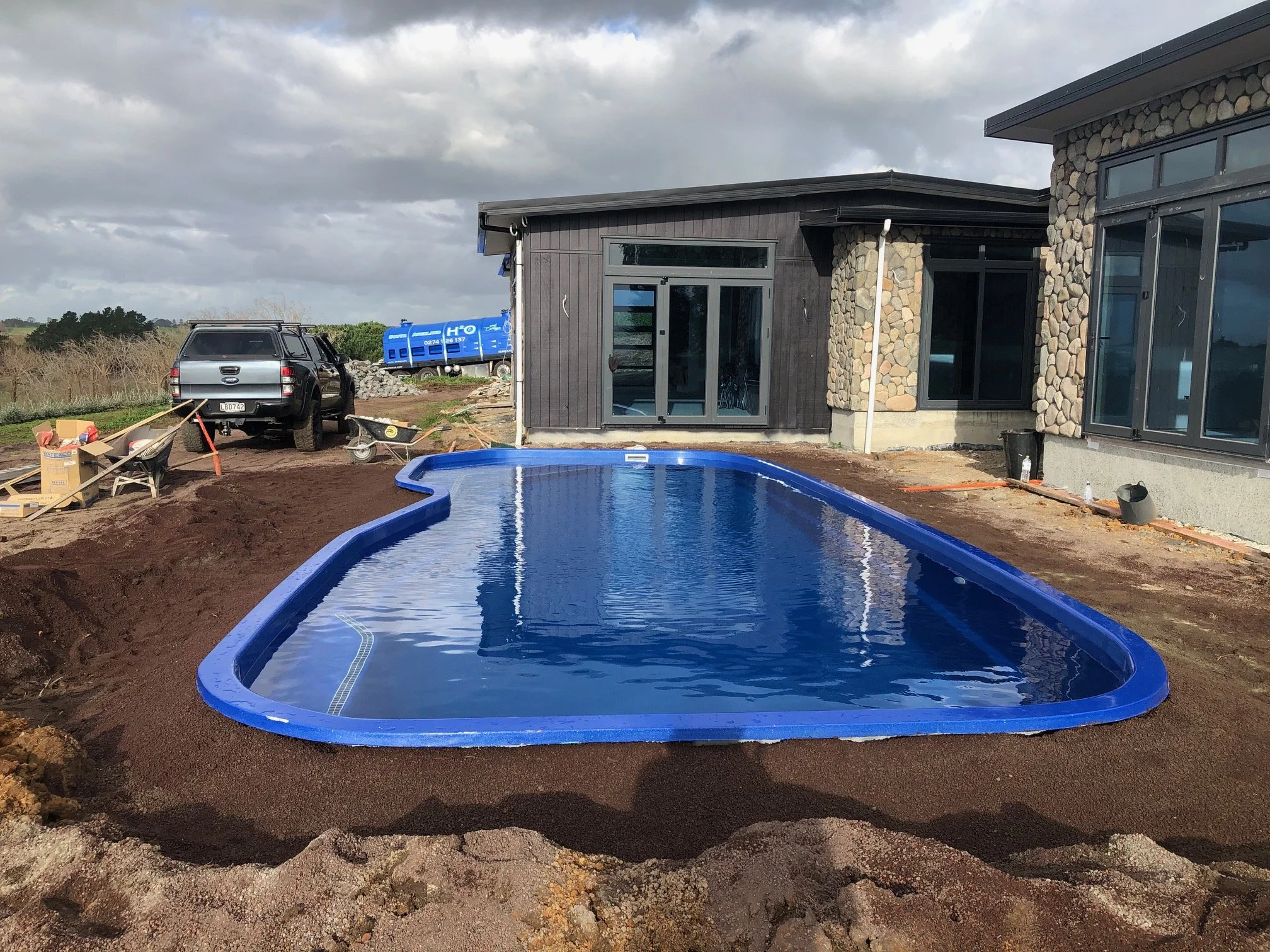 Under construction backyard with a new swimming pool, construction tools, vehicle, and building in the background.