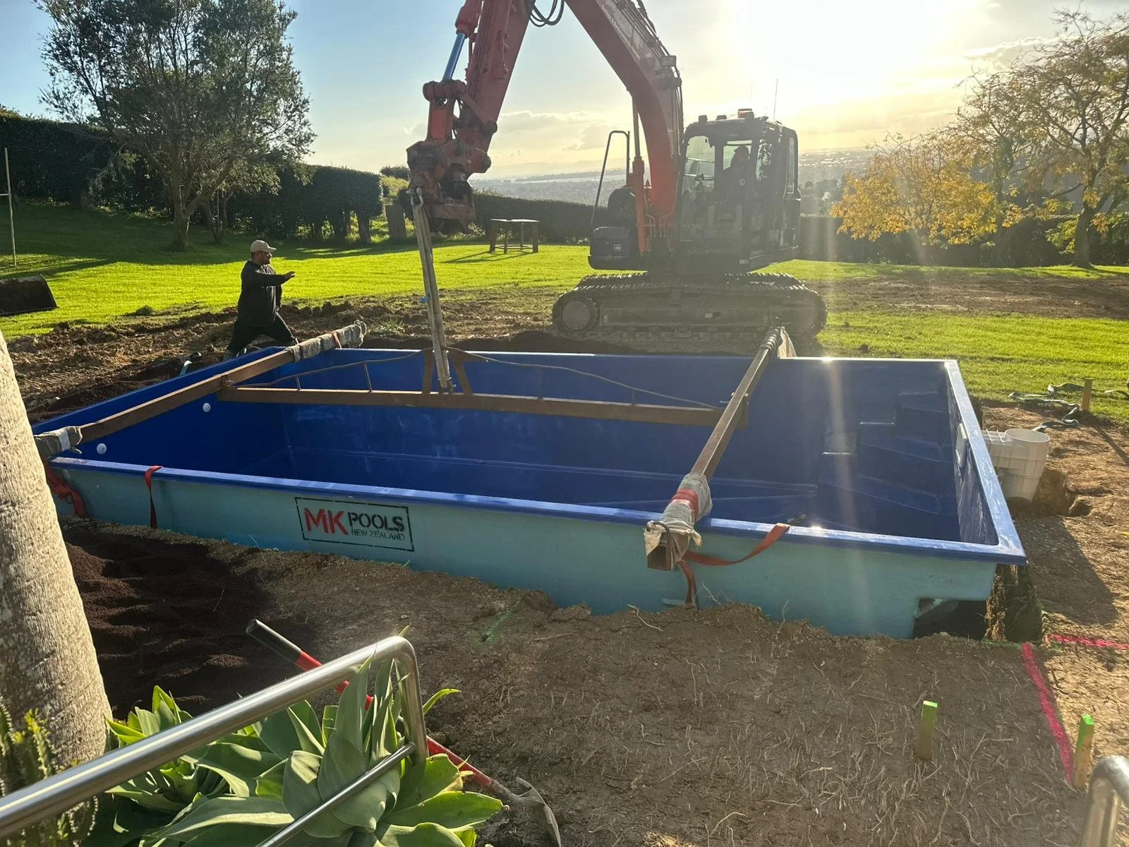 Construction site with a large blue pool labeled 'MK Pools New Zealand,' a person in black walking nearby, and a red excavator in the background on a sunny day with trees and green grass.