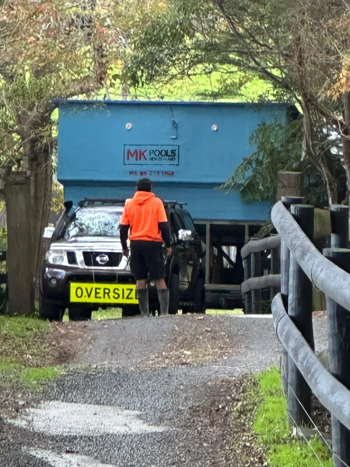 A person wearing an orange hoodie and black shorts standing in front of a large truck with a sign that says 'Oversized.' The truck has a blue container on the back and a logo 'MK Pools' on the front of the container.