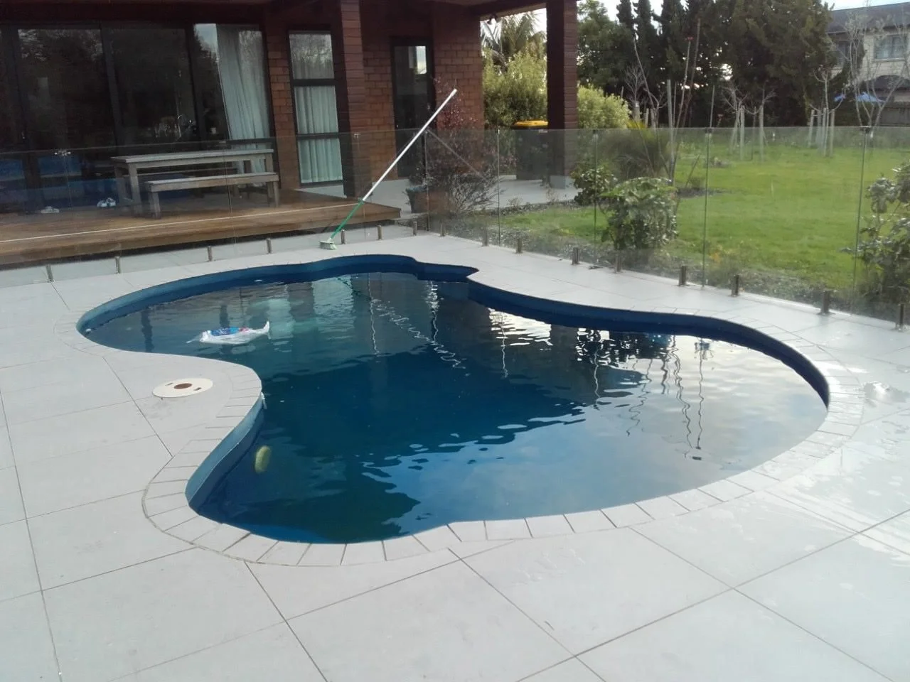 Residential backyard with a kidney-shaped swimming pool, glass fence, and greenery, with a wooden deck and house in the background.