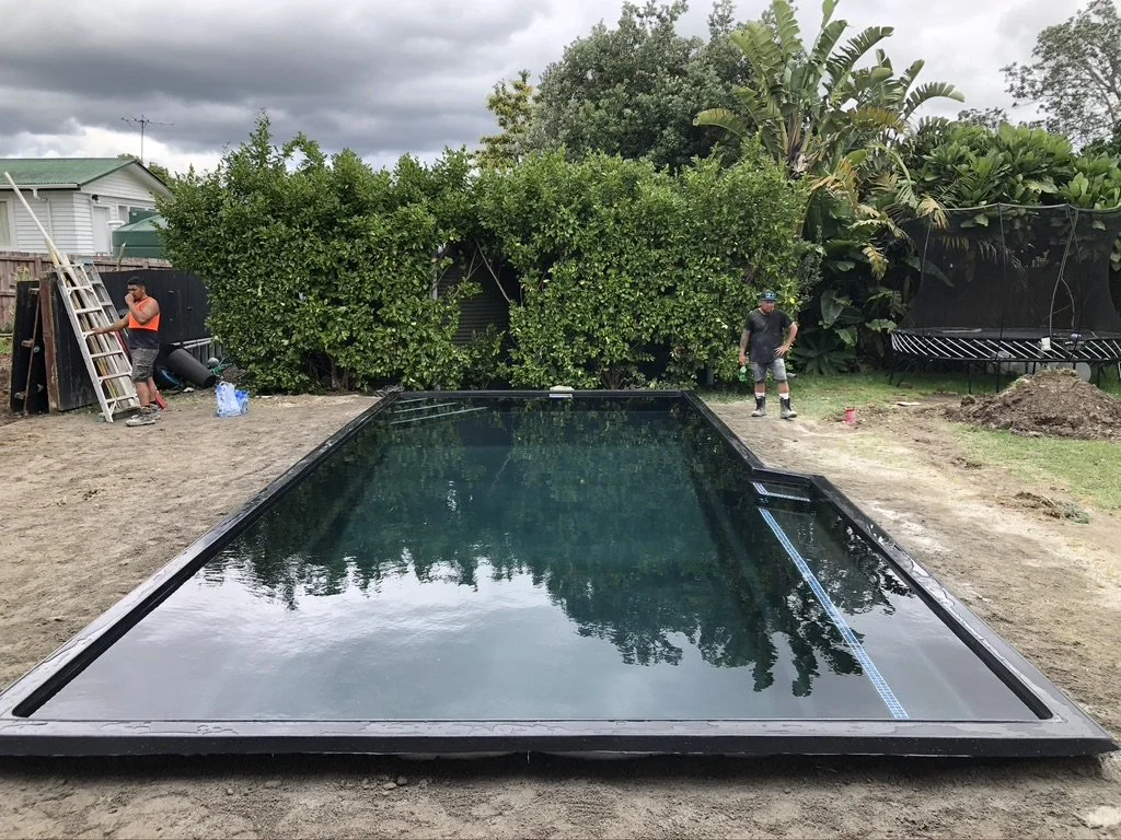 Two men working in a backyard with a large, in-ground swimming pool under construction. One man is on a ladder and the other is standing on the ground, near a trampoline and a pile of dirt. There are bushes, trees, and a house with a green roof in th