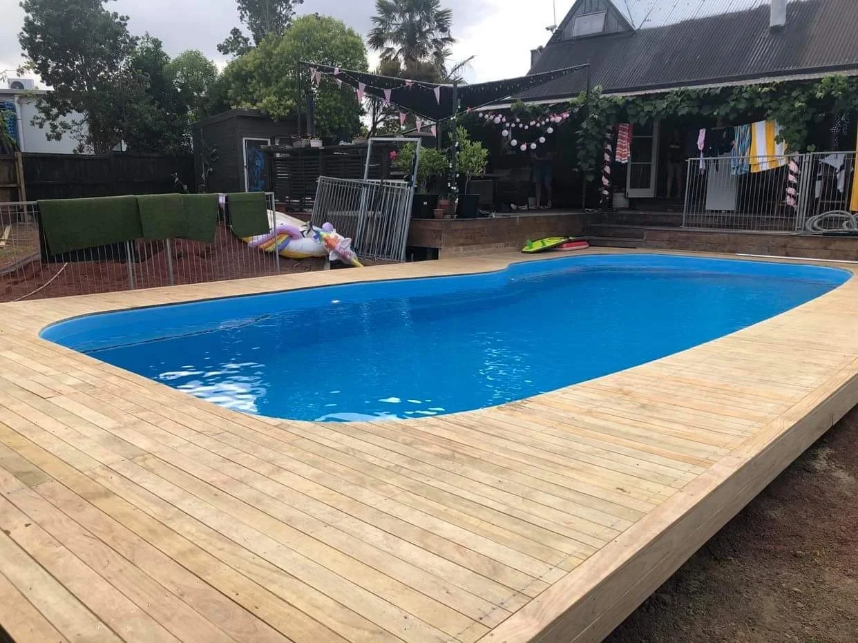 A backyard with an in-ground swimming pool surrounded by a wooden deck, with a house and trees in the background, and pool accessories like floaties and towels on nearby fences and furniture.