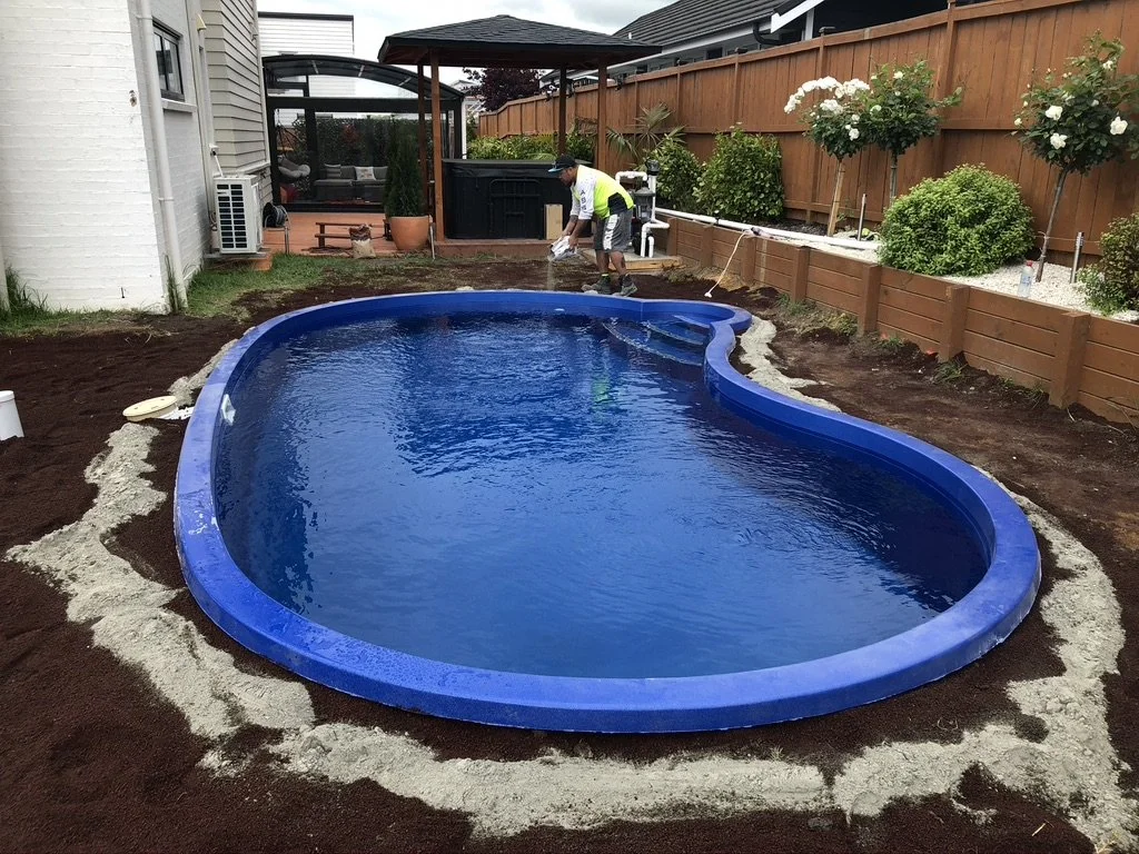 A backyard with a partially installed kidney-shaped swimming pool, surrounded by dirt and a wooden fence. A person is working near the pool with a watering hose, and there are plants and a shed in the background.