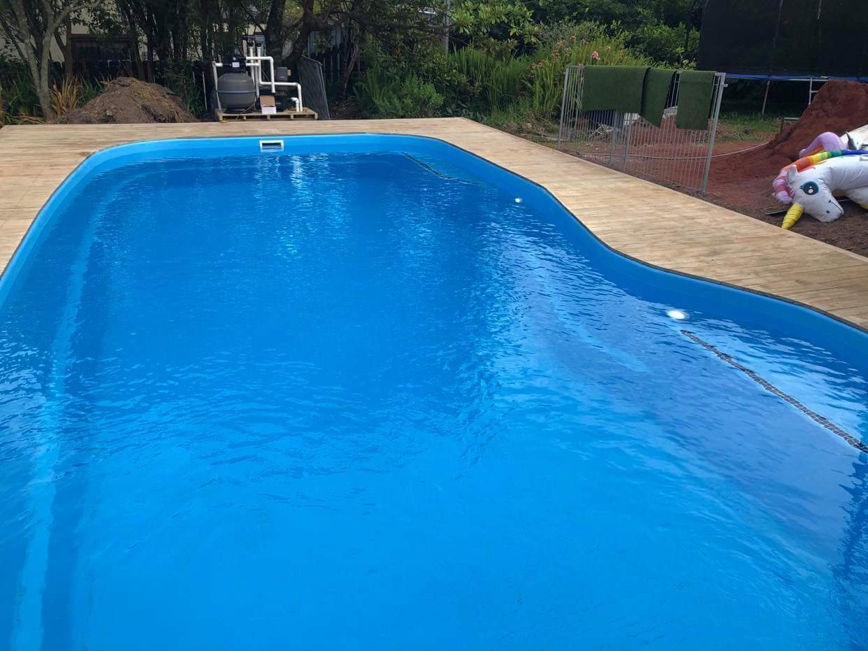 An empty swimming pool with blue water and a wooden deck surrounding it, located outdoors with trees and grass in the background.