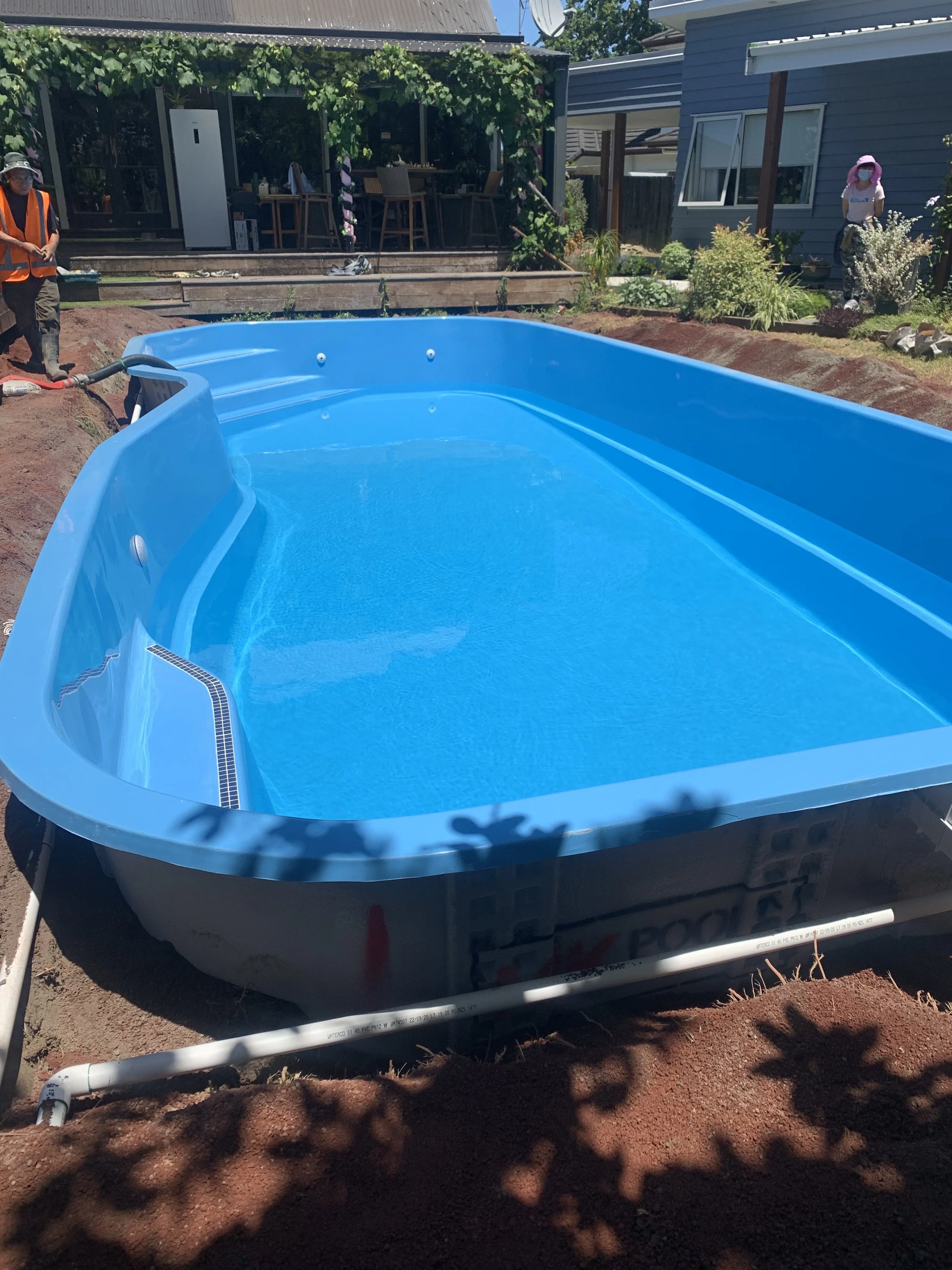 A newly installed above-ground swimming pool in a backyard with brown soil around it, surrounded by a garden with shrubs and a house, under a clear sky.