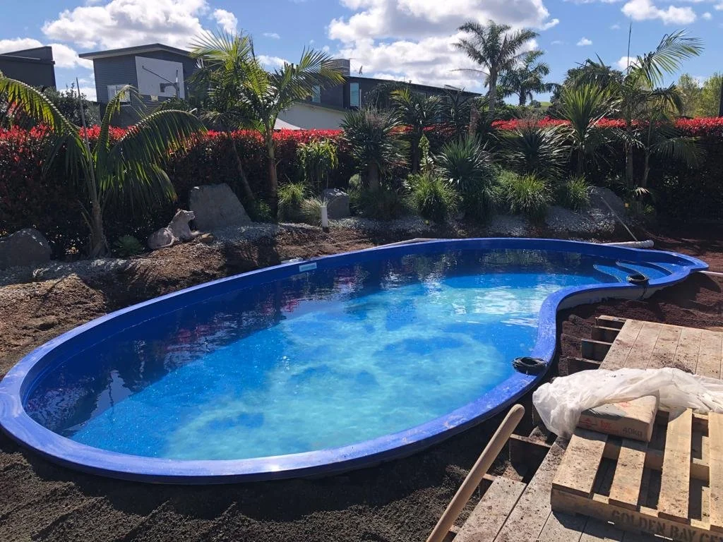 In-ground swimming pool with blue lining, surrounded by tropical plants such as palms, and construction materials like pallets and plastic on the deck, set in a landscaped backyard with modern houses in the background under partly cloudy sky.